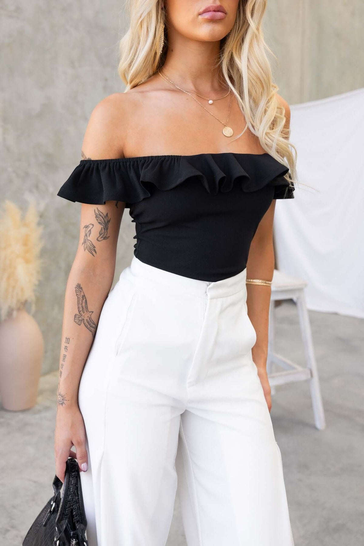 Model wearing a black off-shoulder Bellar bodysuit with ruffles, styled with white pants.