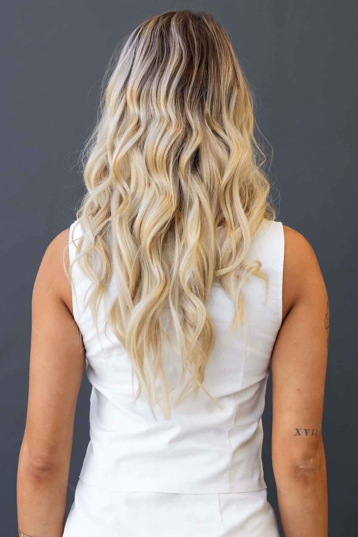 Back view of a woman with wavy blonde hair wearing a white sleeveless top against a gray background.