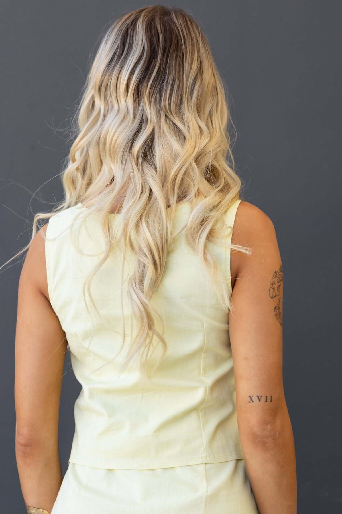 Back view of a woman wearing a light yellow Pamela Top with a button-up front and flowing blonde hair.