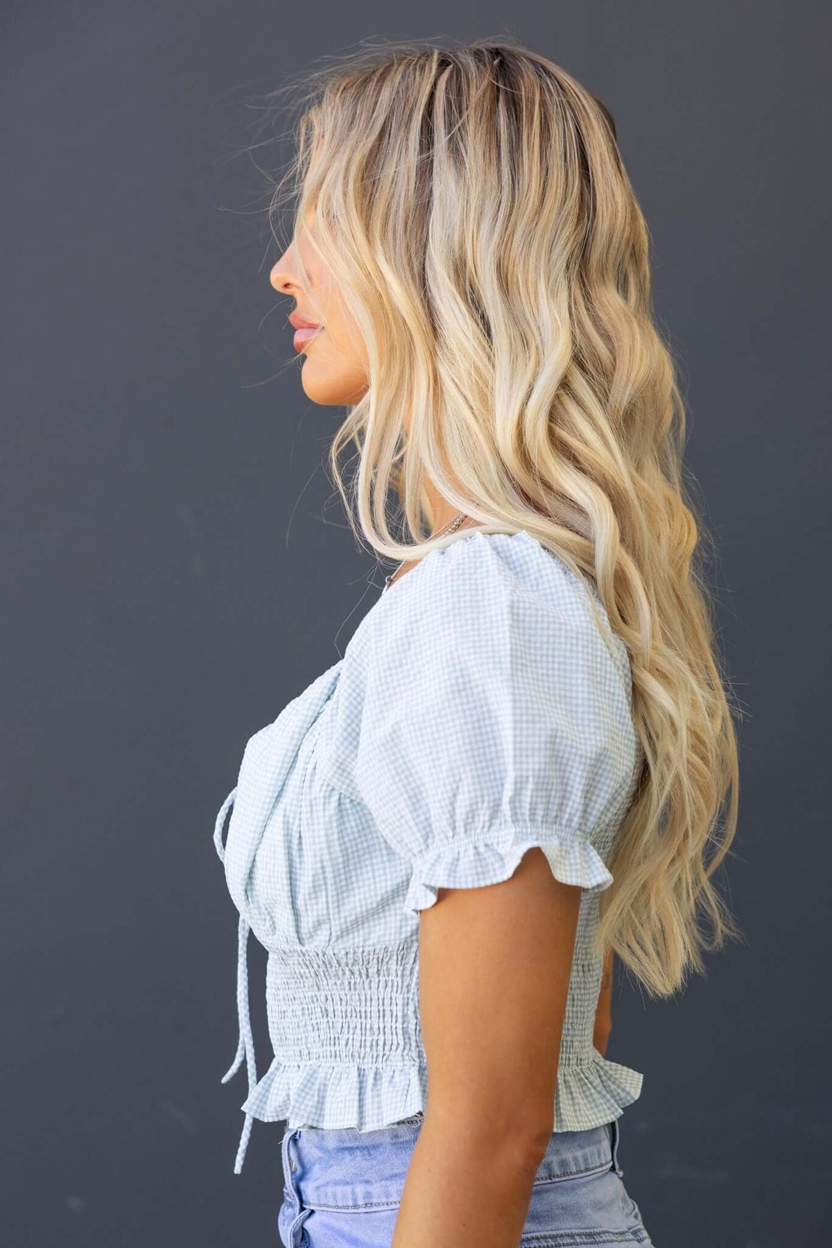 Profile view of a woman wearing a blue and white checked Sonnie Top with a tie at the bust and ruffled sleeves.