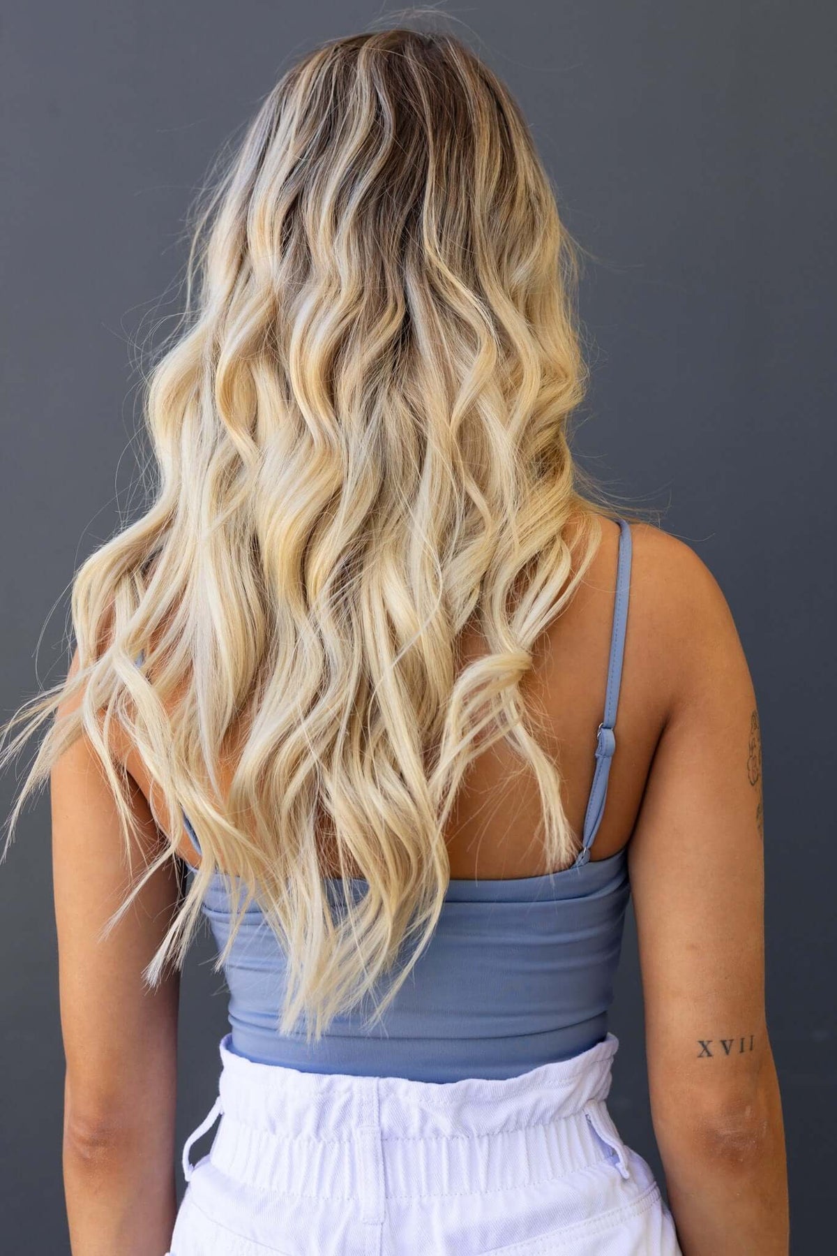Back view of a woman wearing a gray bodysuit with wavy long blonde hair, showcasing adjustable straps.