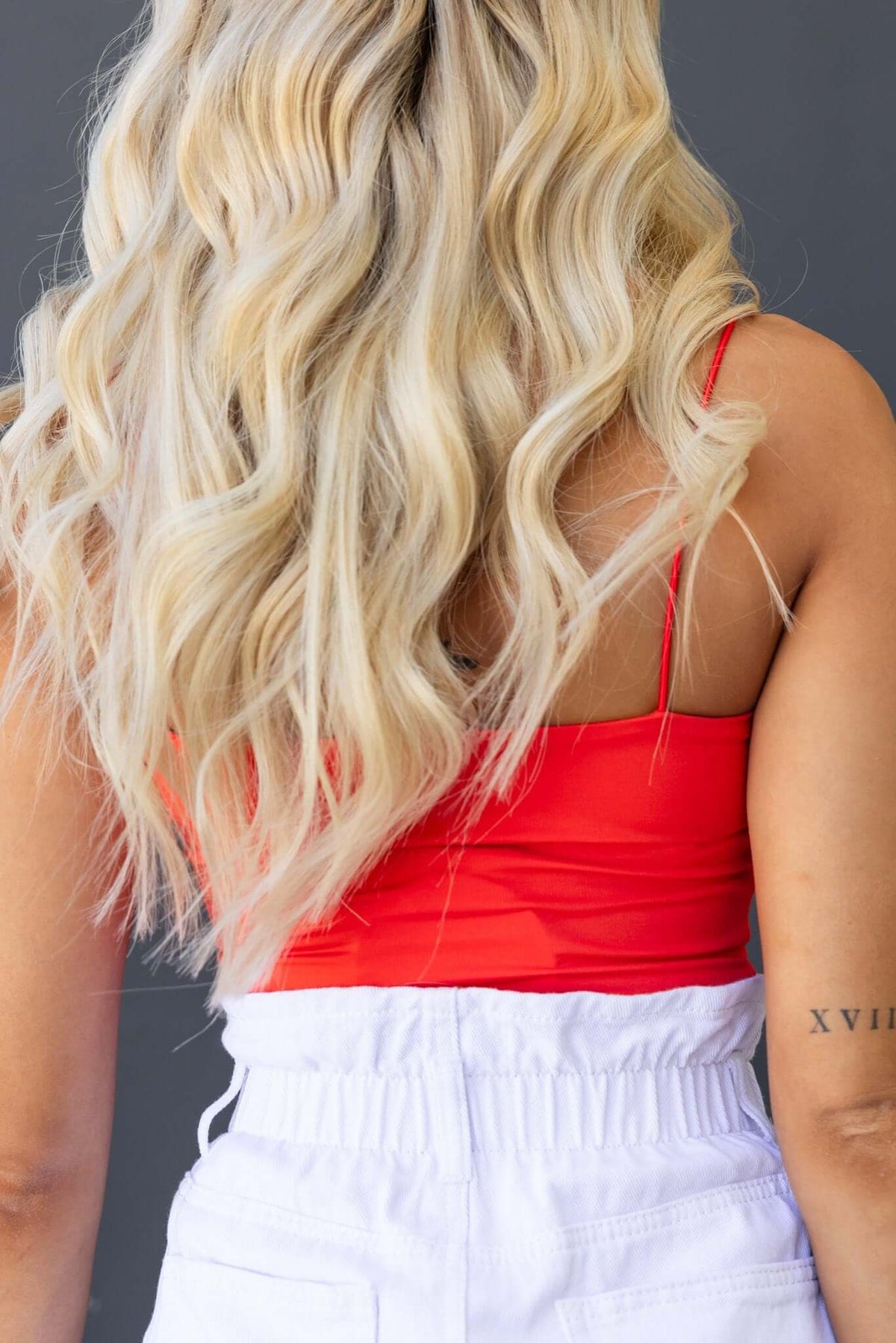 Back view of a woman in a ruby bodysuit with spaghetti straps, styled with white pants and long, wavy blonde hair.