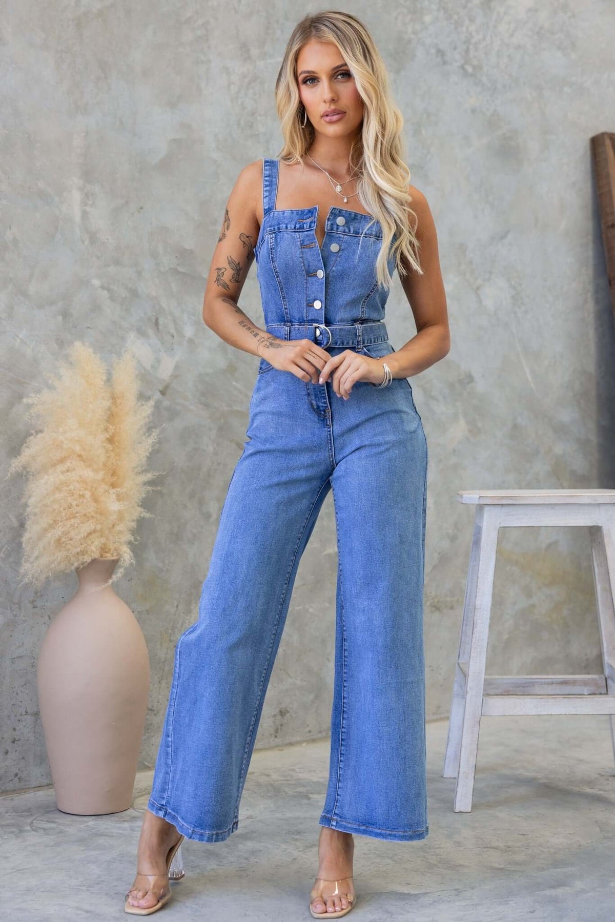 Tamara models the Josey Denim Jumpsuit featuring adjustable straps, button-up front, and a removable belt.