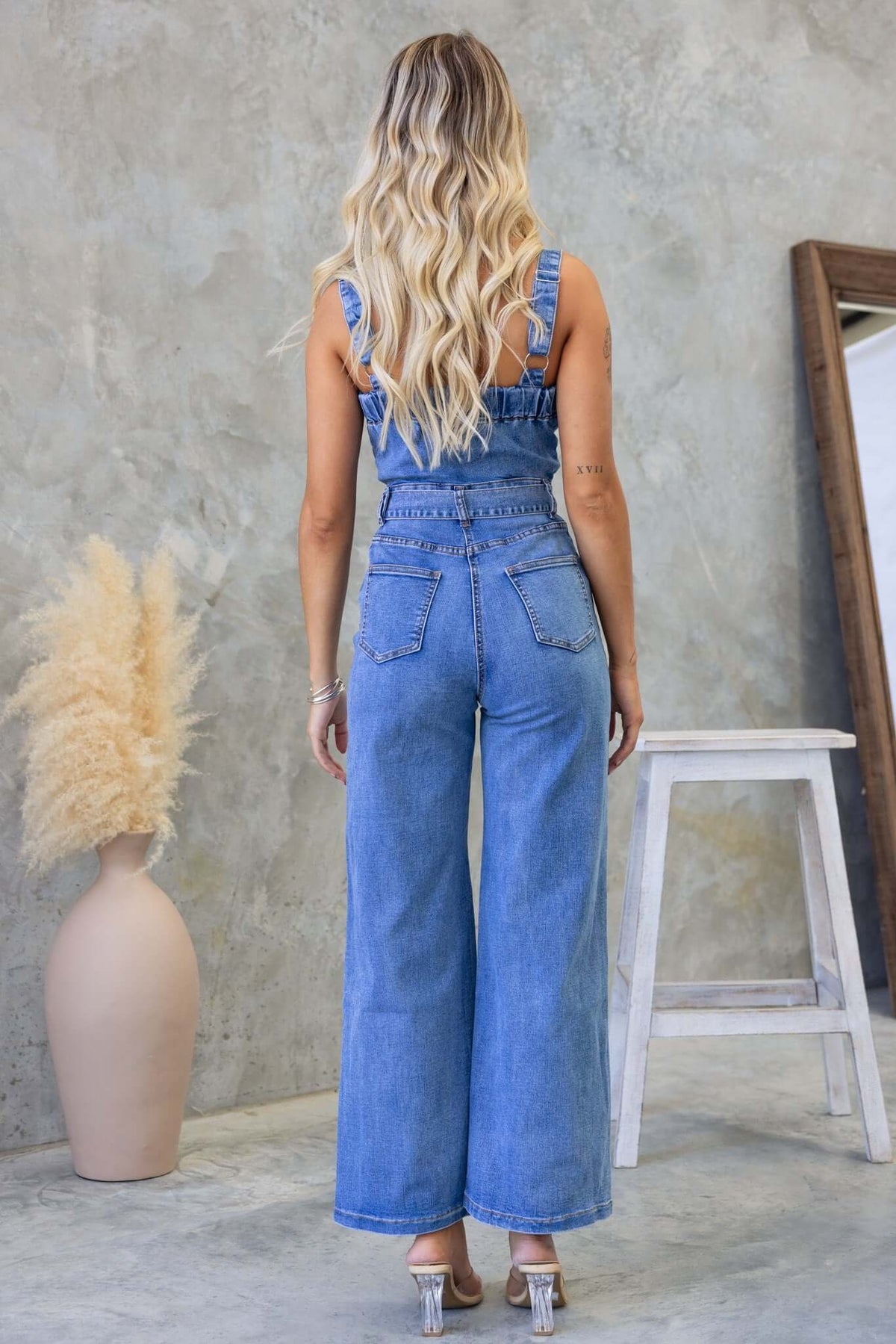 Back view of a woman in a Josey Denim Jumpsuit, featuring adjustable straps and wide-leg design, set in a modern interior.