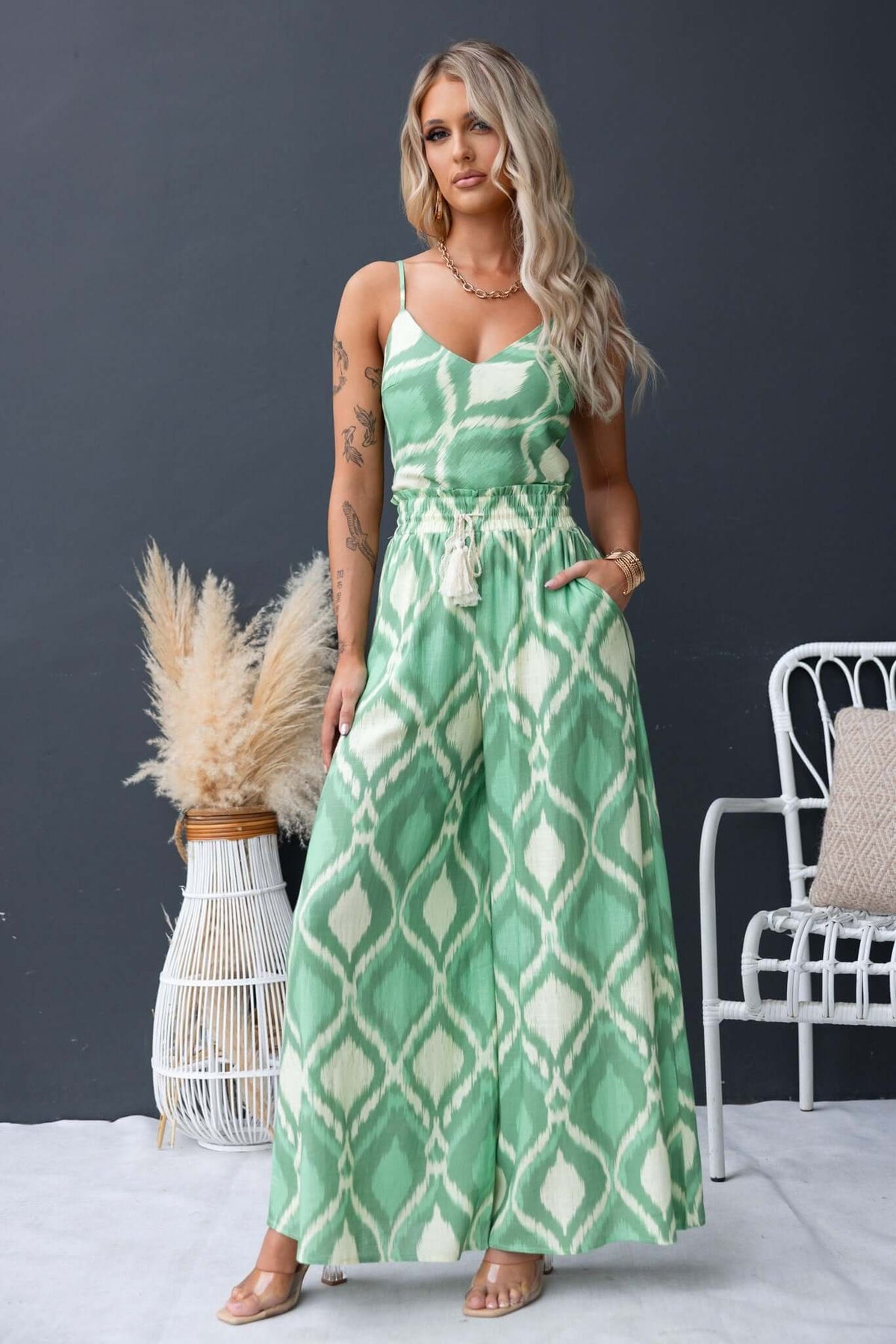 Model wearing a green patterned jumpsuit with spaghetti straps and a drawstring waist, set against a dark background.