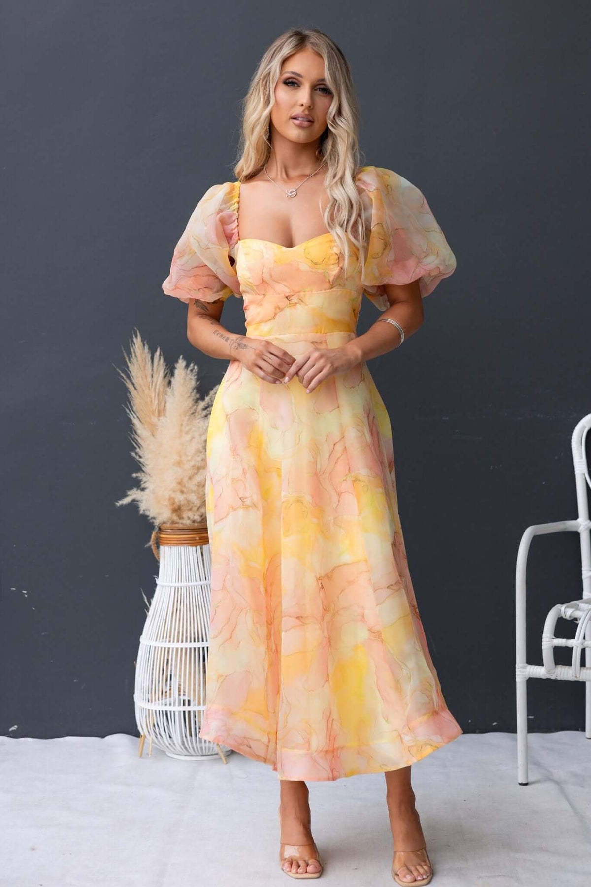 Model wearing a Nerine Maxi Dress in pastel yellow with puff sleeves and floral print, styled with a boho decor.