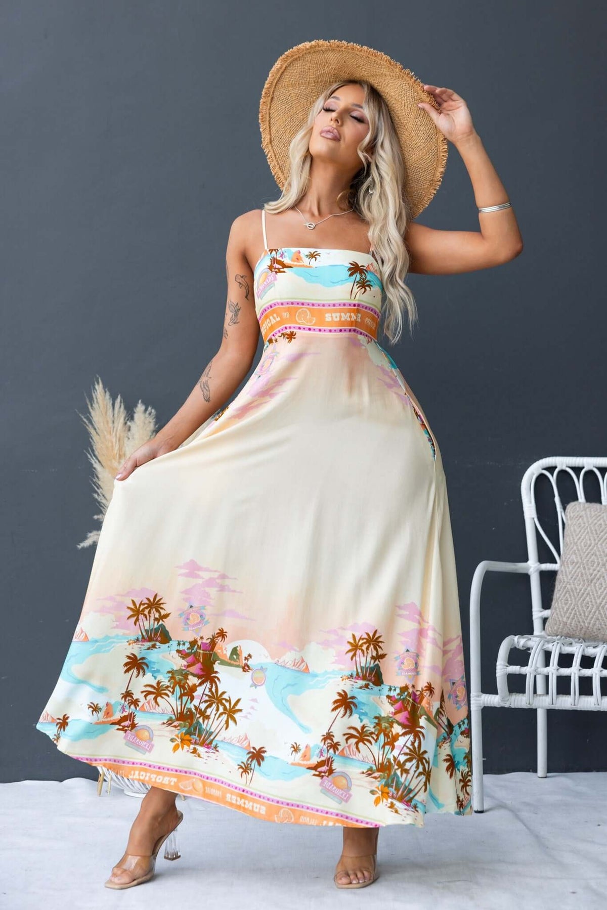 Model wearing Salilah Maxi Dress with floral design, thin straps, and a straw hat, posing against a gray background.