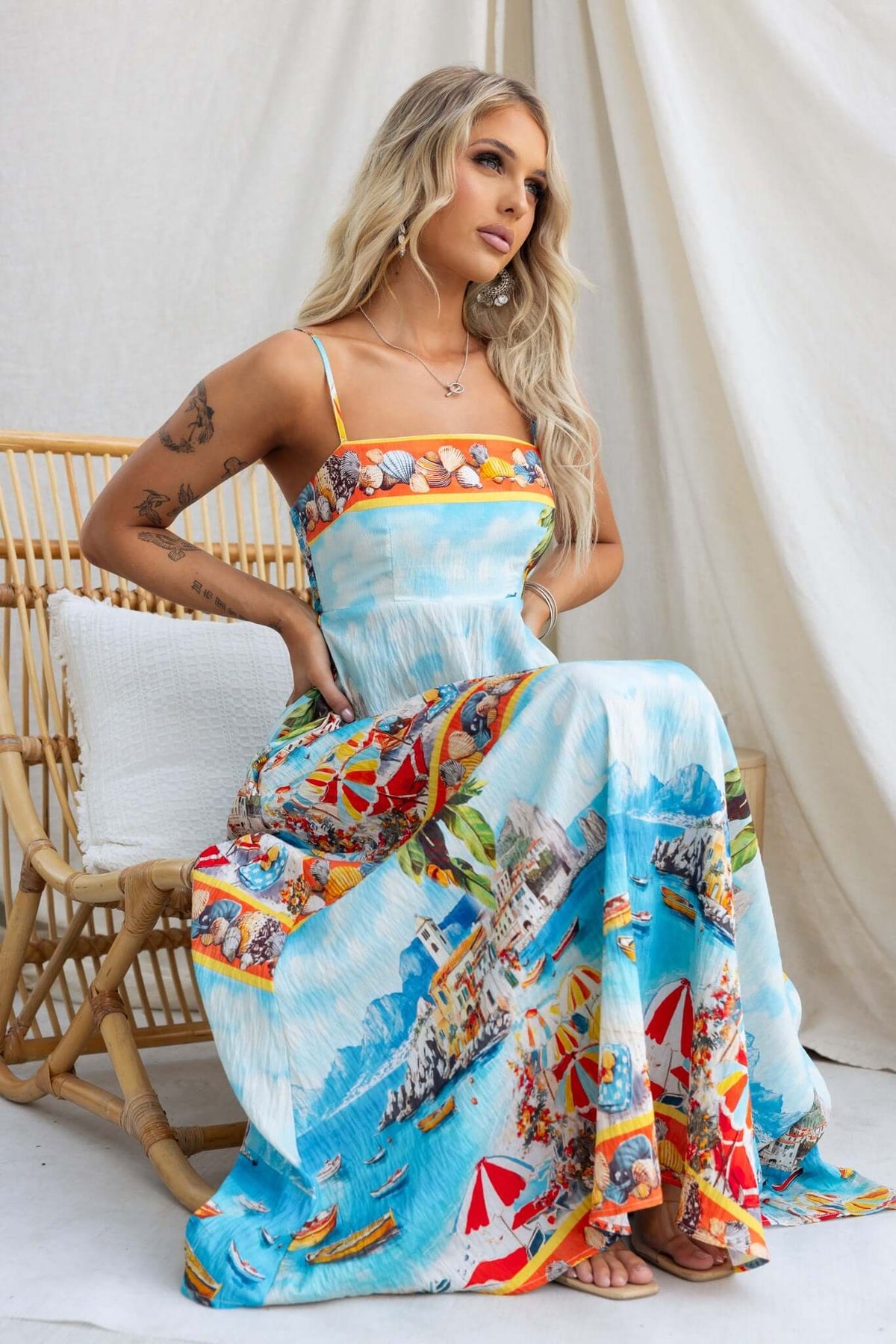 Model wearing Salilah Maxi Dress with colorful beach print, thin adjustable straps, and pockets, seated against a neutral backdrop.