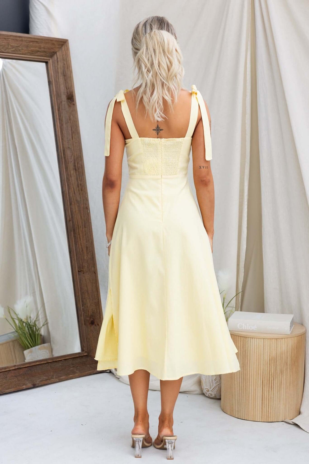 Back view of a woman in a yellow Palomia Midi Dress with adjustable tie straps, shown in a soft interior setting.