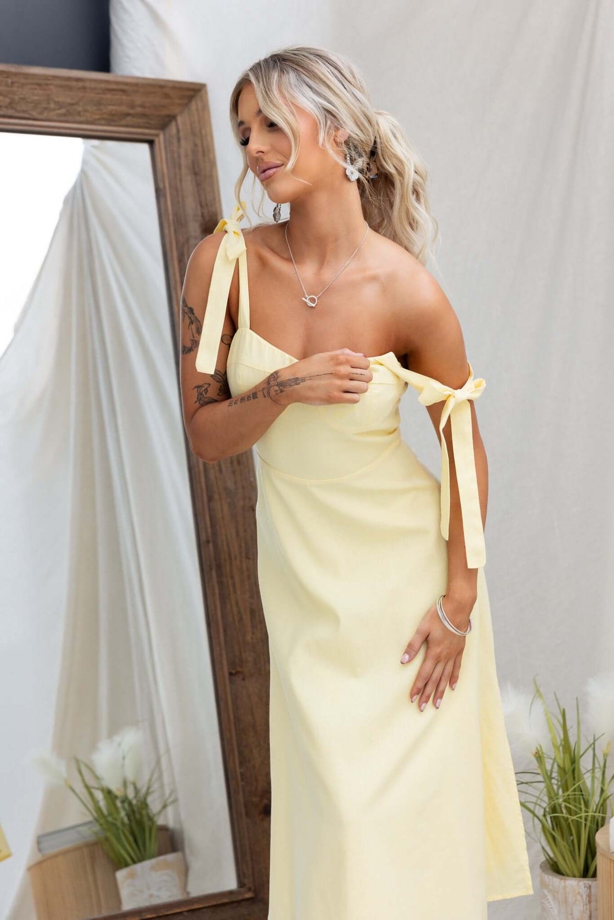 Woman wearing Palomia Midi Dress in soft yellow with adjustable tie straps and side split, mirrored backdrop.