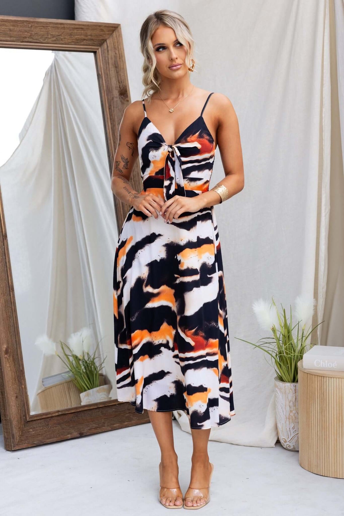 Model wearing Cecilee Midi Dress with adjustable straps and tie at bust, featuring a colorful pattern in front of a mirror.