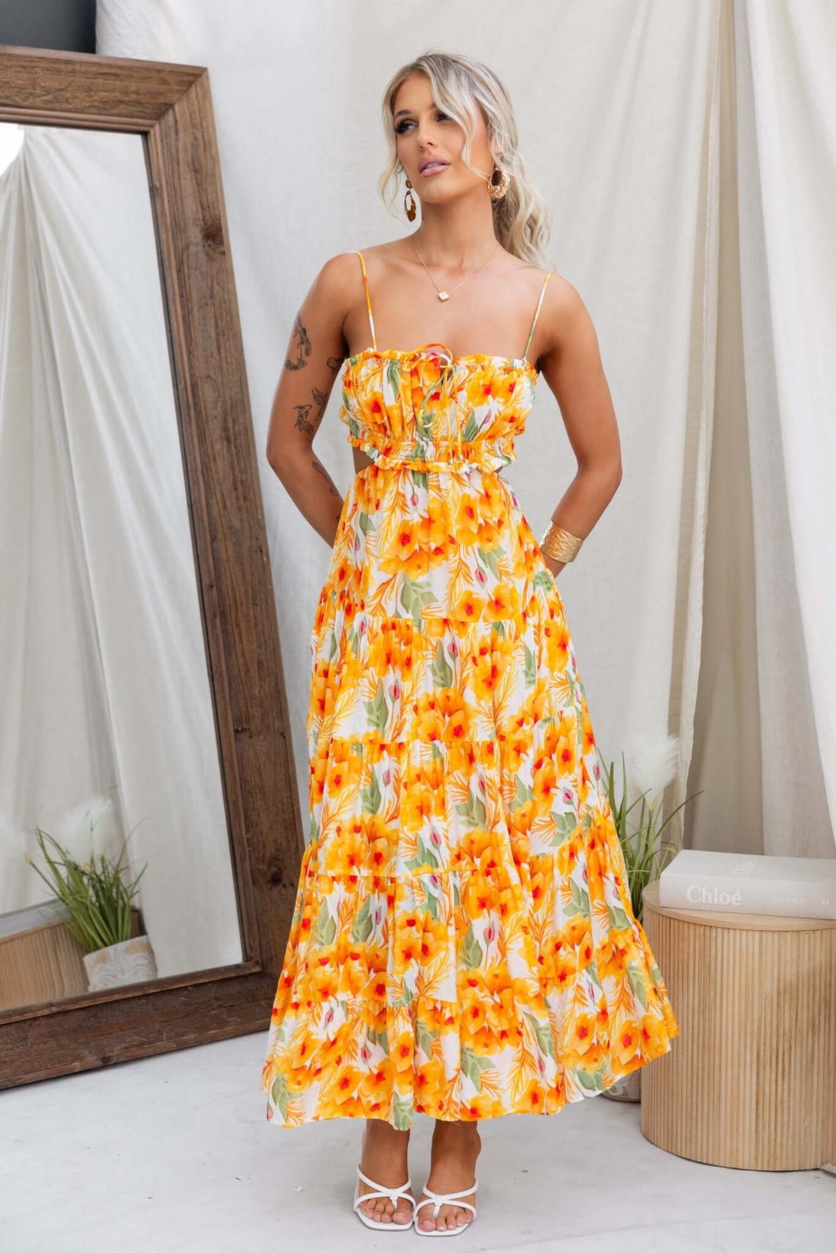 Model wearing a vibrant orange floral Kohana maxi dress with adjustable straps and a tie at the back, styled in a chic indoor setting.
