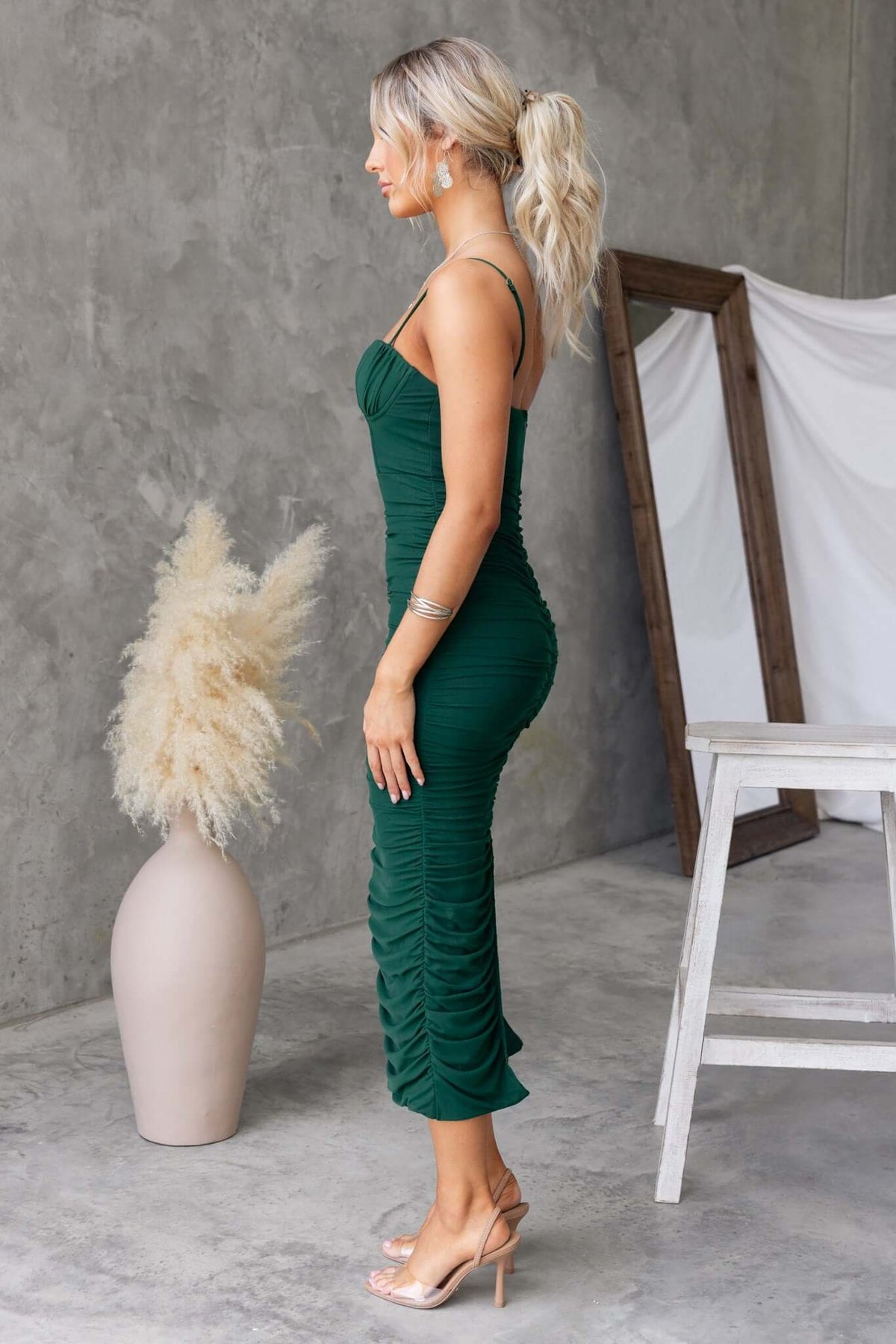 Model in a green midi dress with padded bust, adjustable straps, and back split on a modern concrete backdrop.