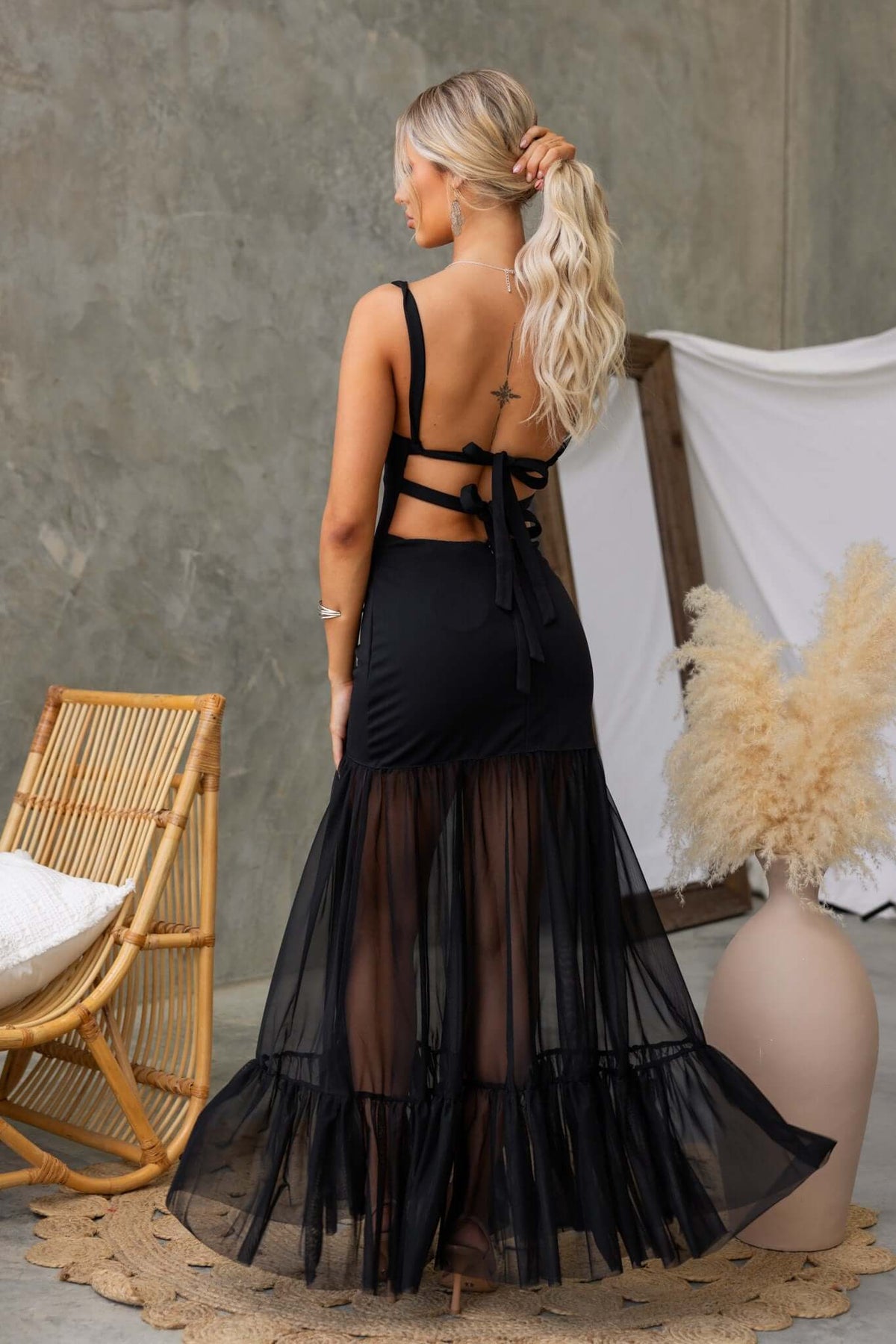 Back view of a woman in a black Nephrite maxi dress with tie straps and sheer mesh skirt, styled in a modern setting.