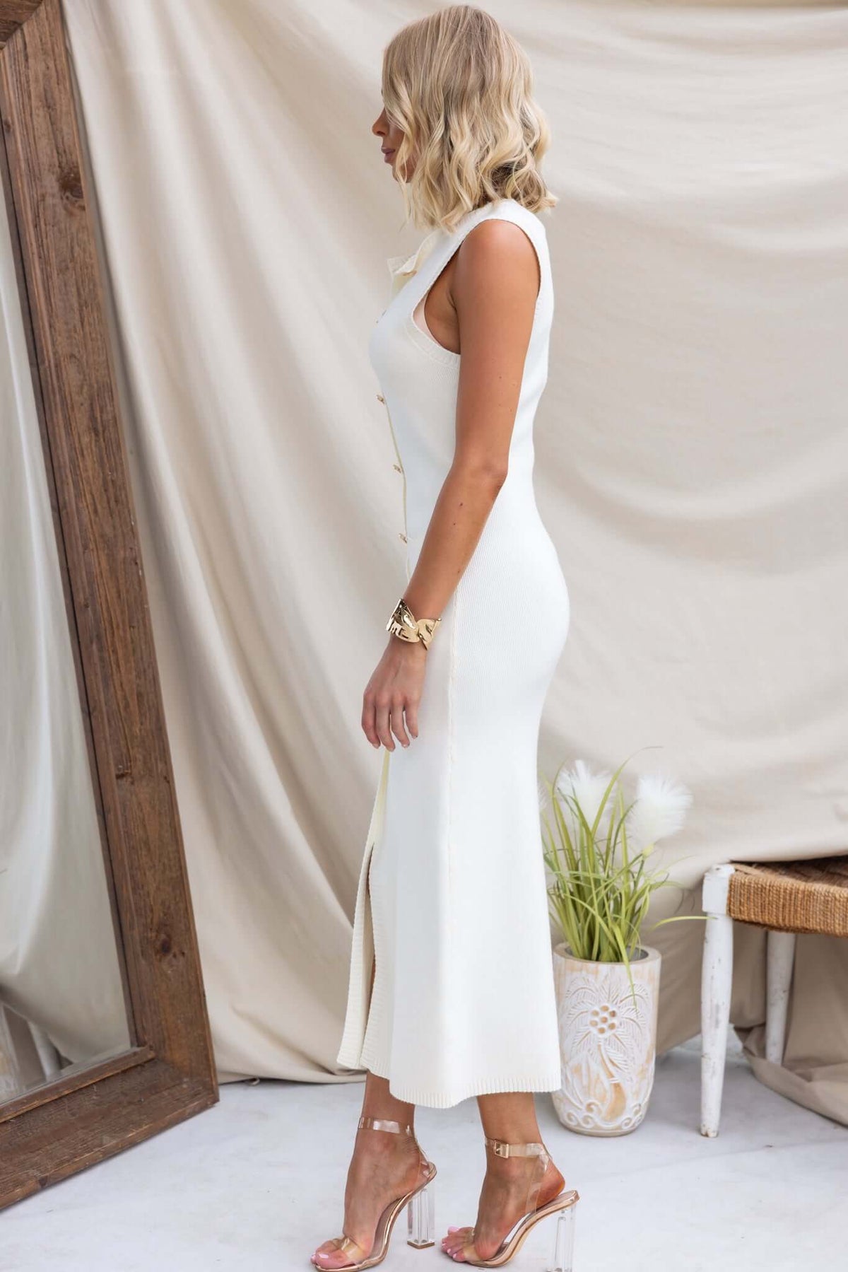 Side view of Bari Maxi Dress in cream with high neckline, faux buttons, split front, and pockets, styled with heels.
