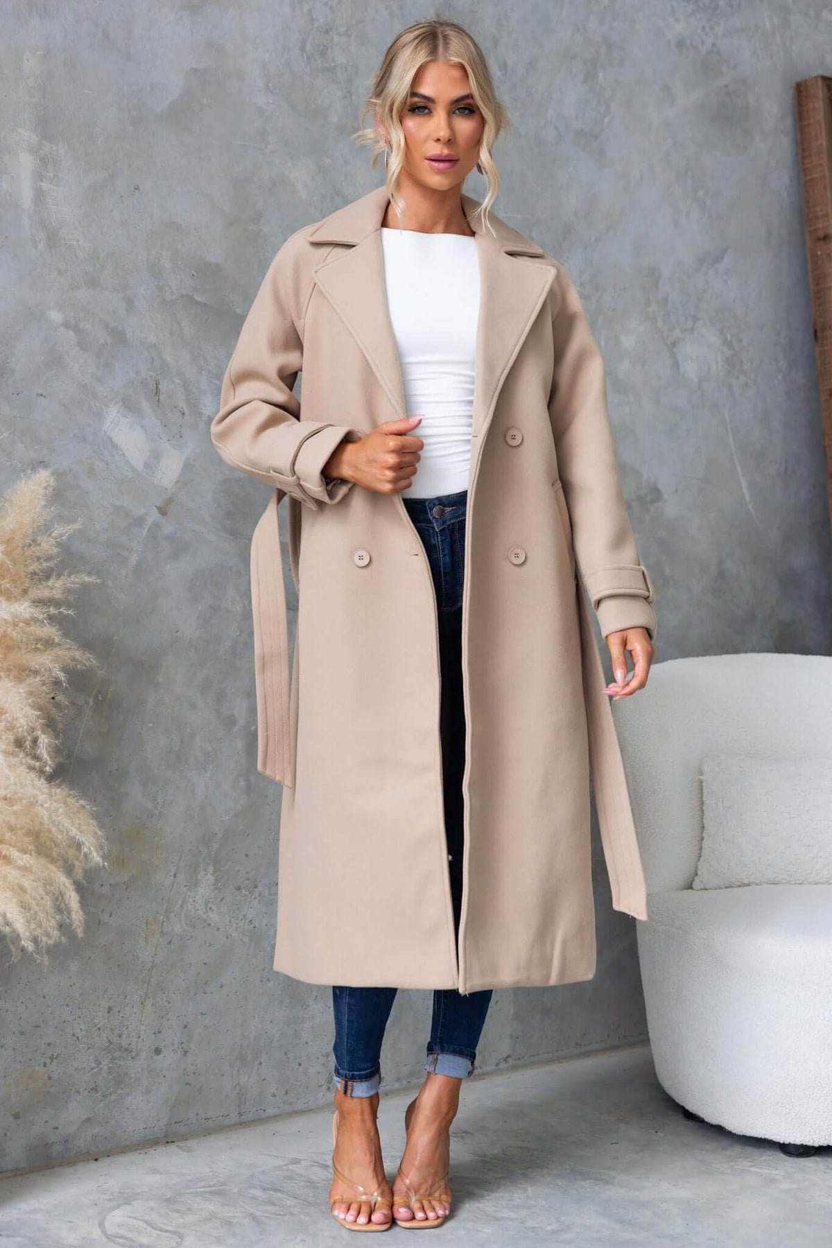 Model wearing a longline beige Farina coat with collar, button-up front, and removable fabric tie, styled with jeans.