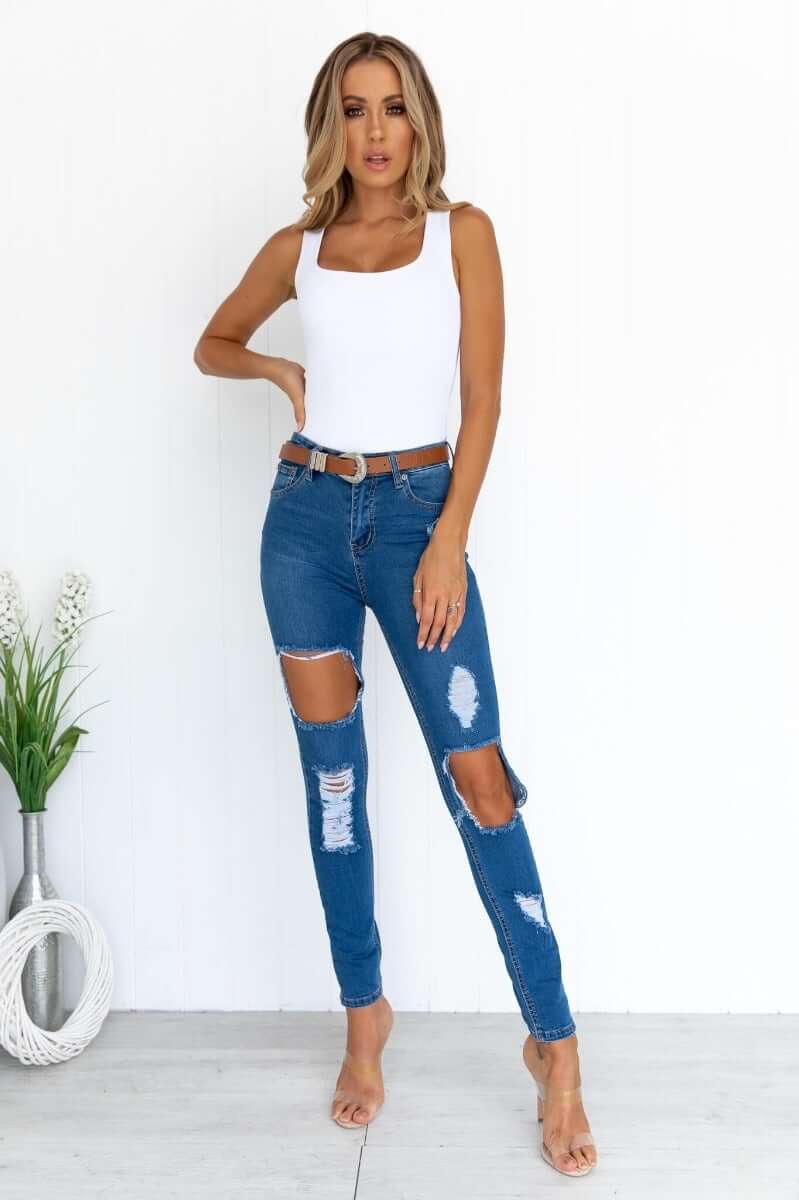 Woman wearing Glory Jeans, distressed denim with rips, paired with a white tank top and heels, standing against a simple background.