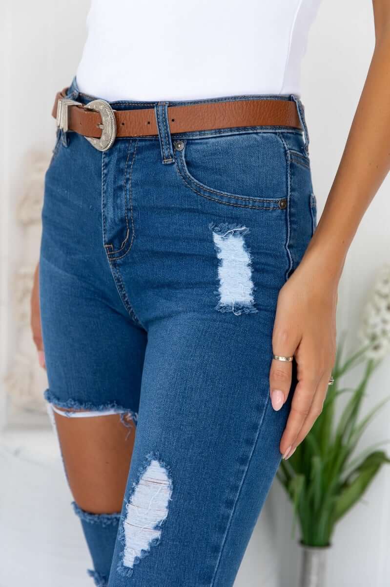 Close-up of distressed Glory Jeans with pockets and rip details, paired with a brown belt, showcasing stretch denim.