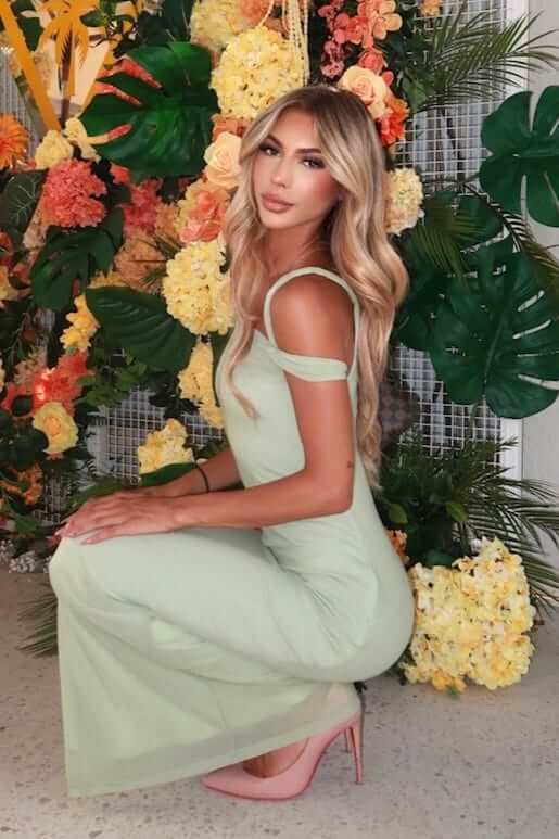 Model wearing a light green Granada maxi dress with twist straps, posing in front of a floral backdrop.