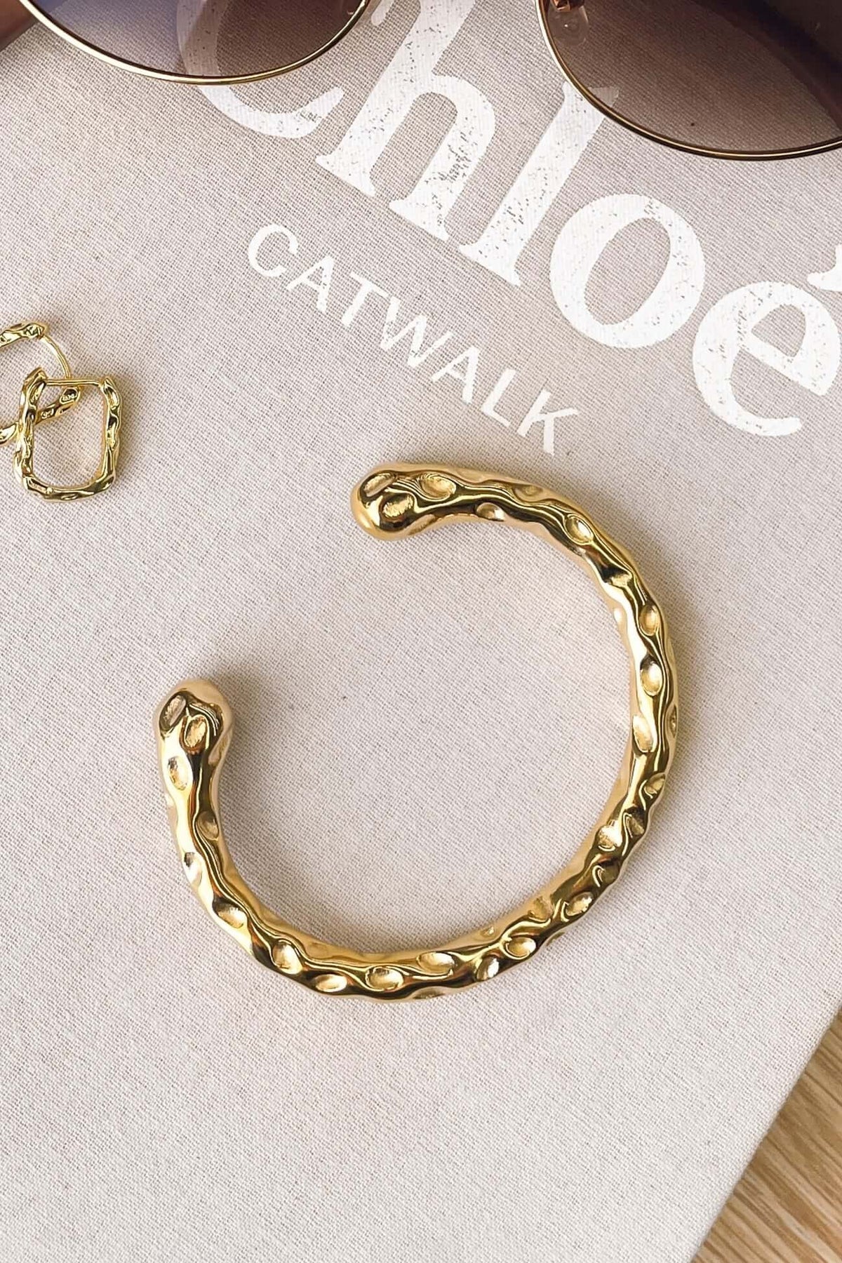 Ever After Bracelet in textured gold, open cuff style, displayed on a fashion magazine with sunglasses.
