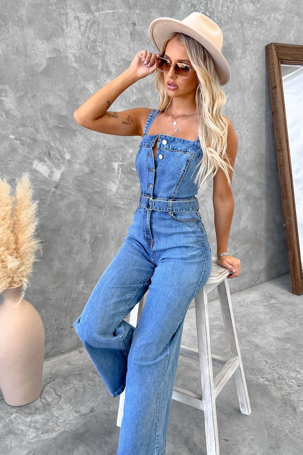 Stylish woman wearing a Josey Denim Jumpsuit with adjustable straps and a removable belt, seated on a stool.