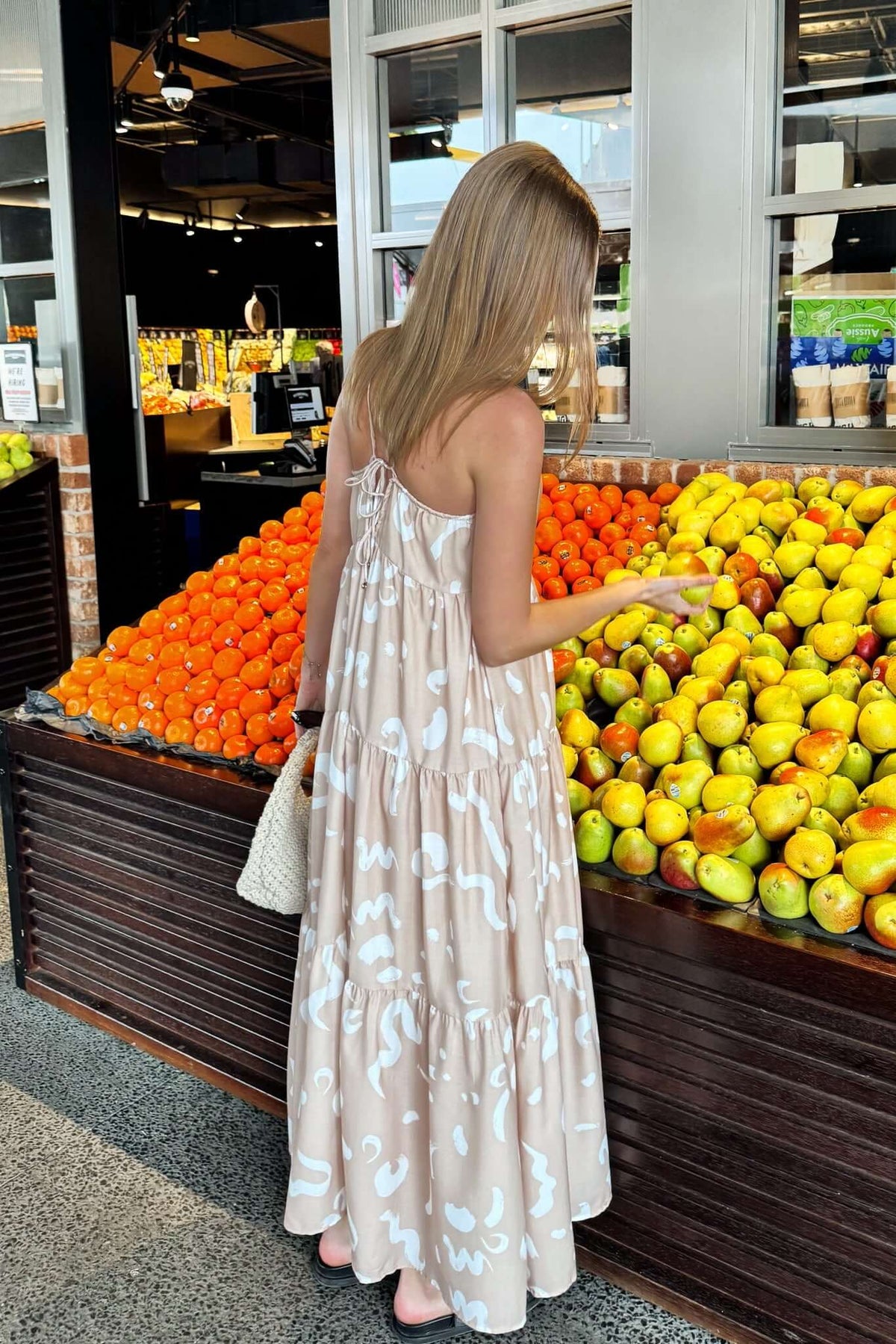 Woman standing in a Udelia maxi dress with square neckline by a fruit stand filled with fresh oranges and lemons.