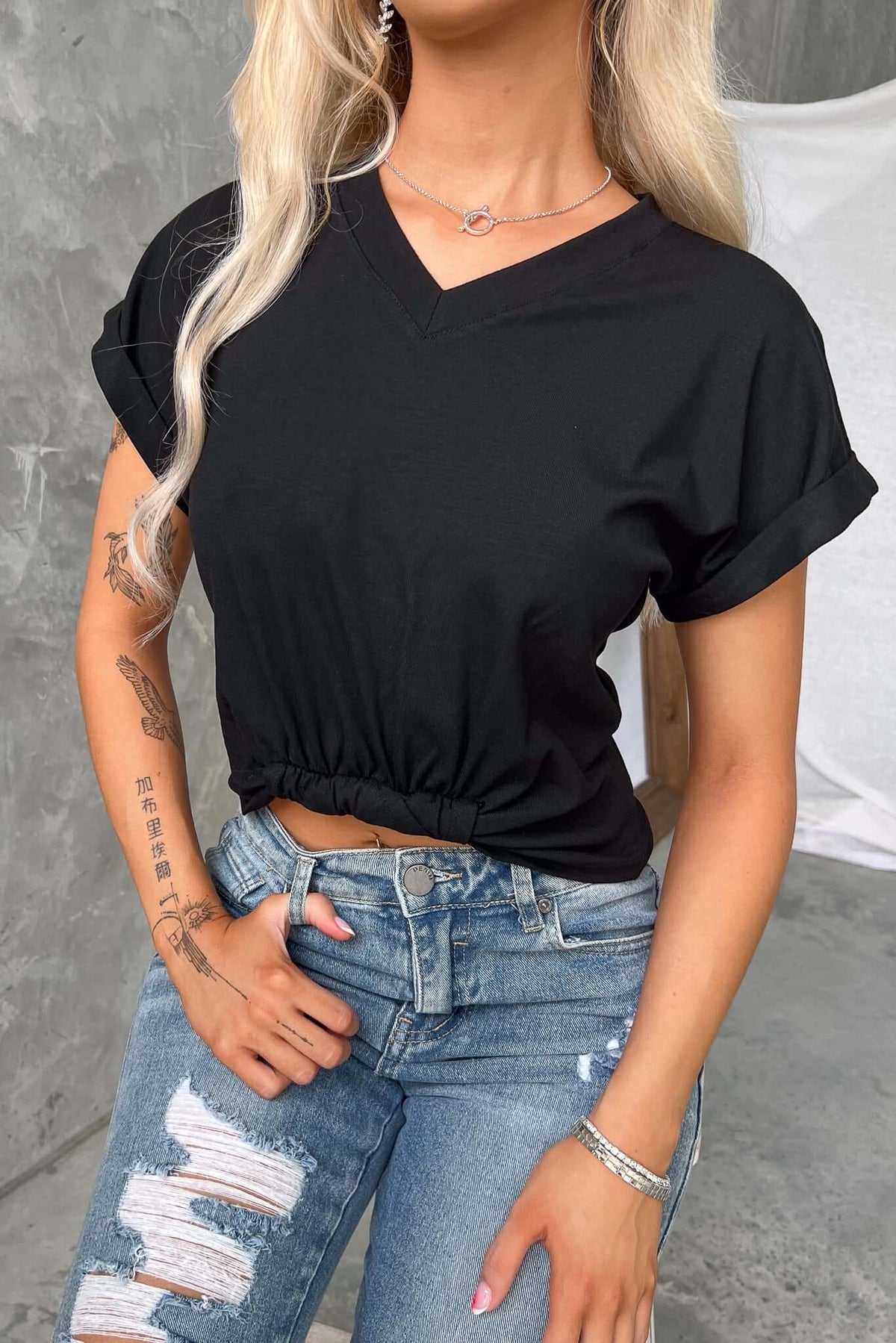 Black V-neck tee with pin roll cuffs and twist at hem, styled with distressed jeans and a casual look.