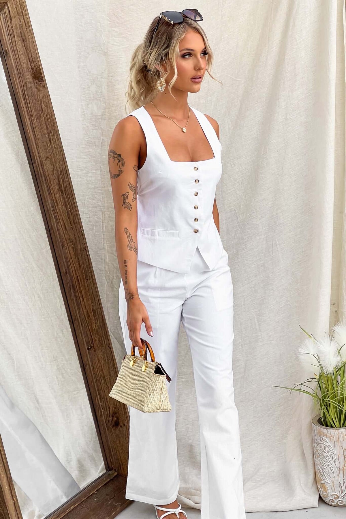 Model wearing a white sleeveless top and matching pants, holding a small handbag, posing against a neutral backdrop.