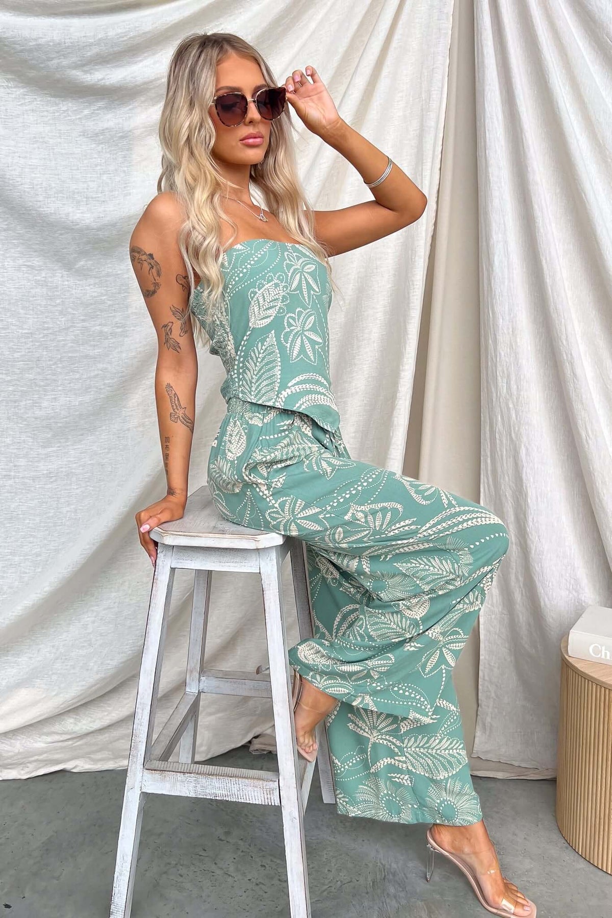 Model in a strapless Lasan Set featuring a printed top and non-stretch pants, sitting on a stool with sunglasses.
