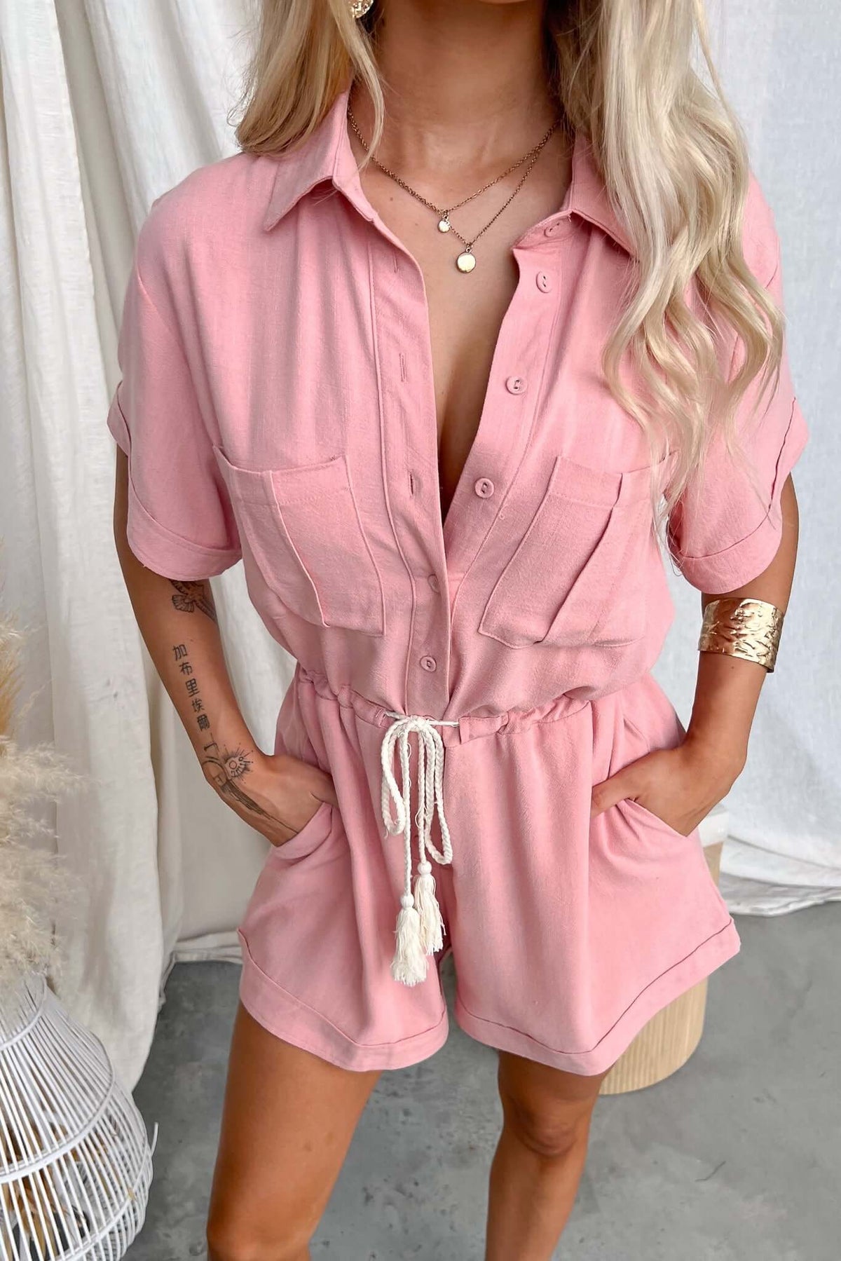 Woman wearing a pink button-up barre playsuit with collar, pockets, and drawstring waist, styled casually.