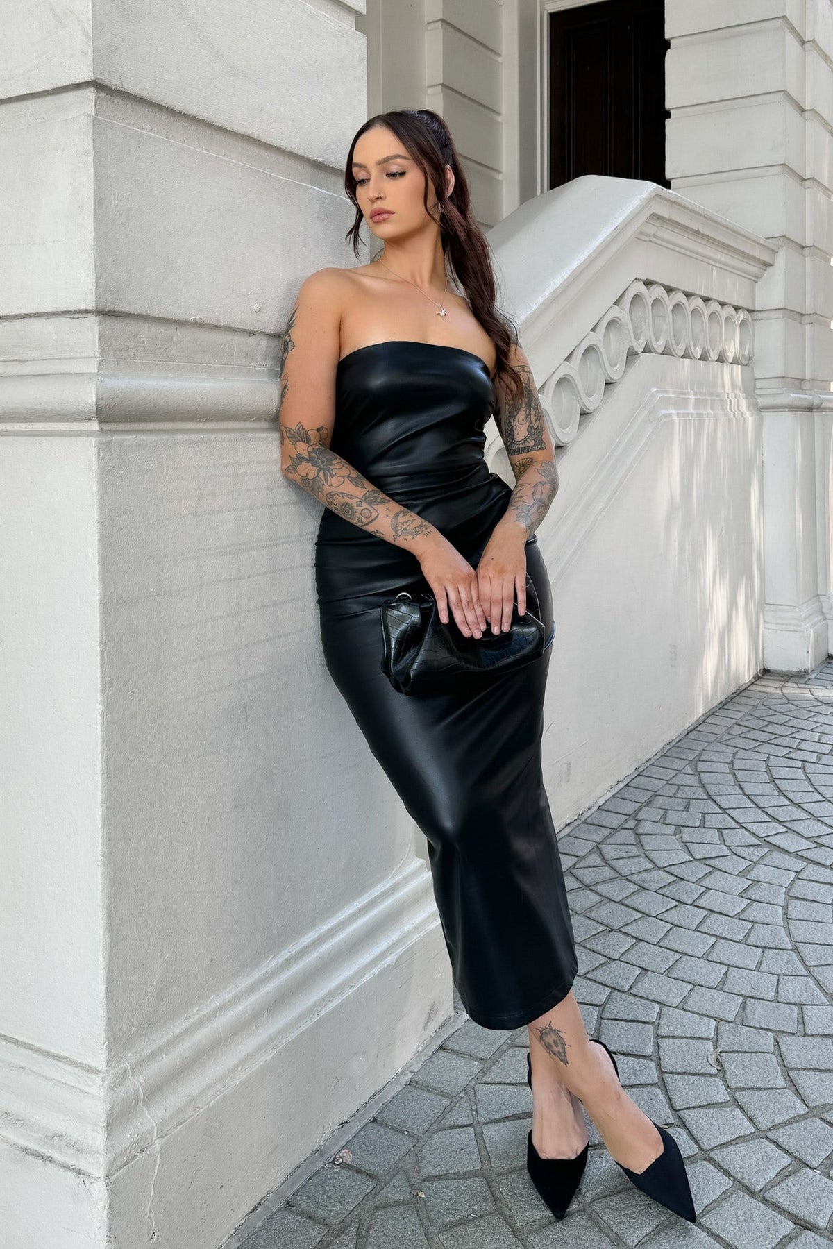 Model in a strapless black midi dress with faux leather material, standing on a stylish urban sidewalk.
