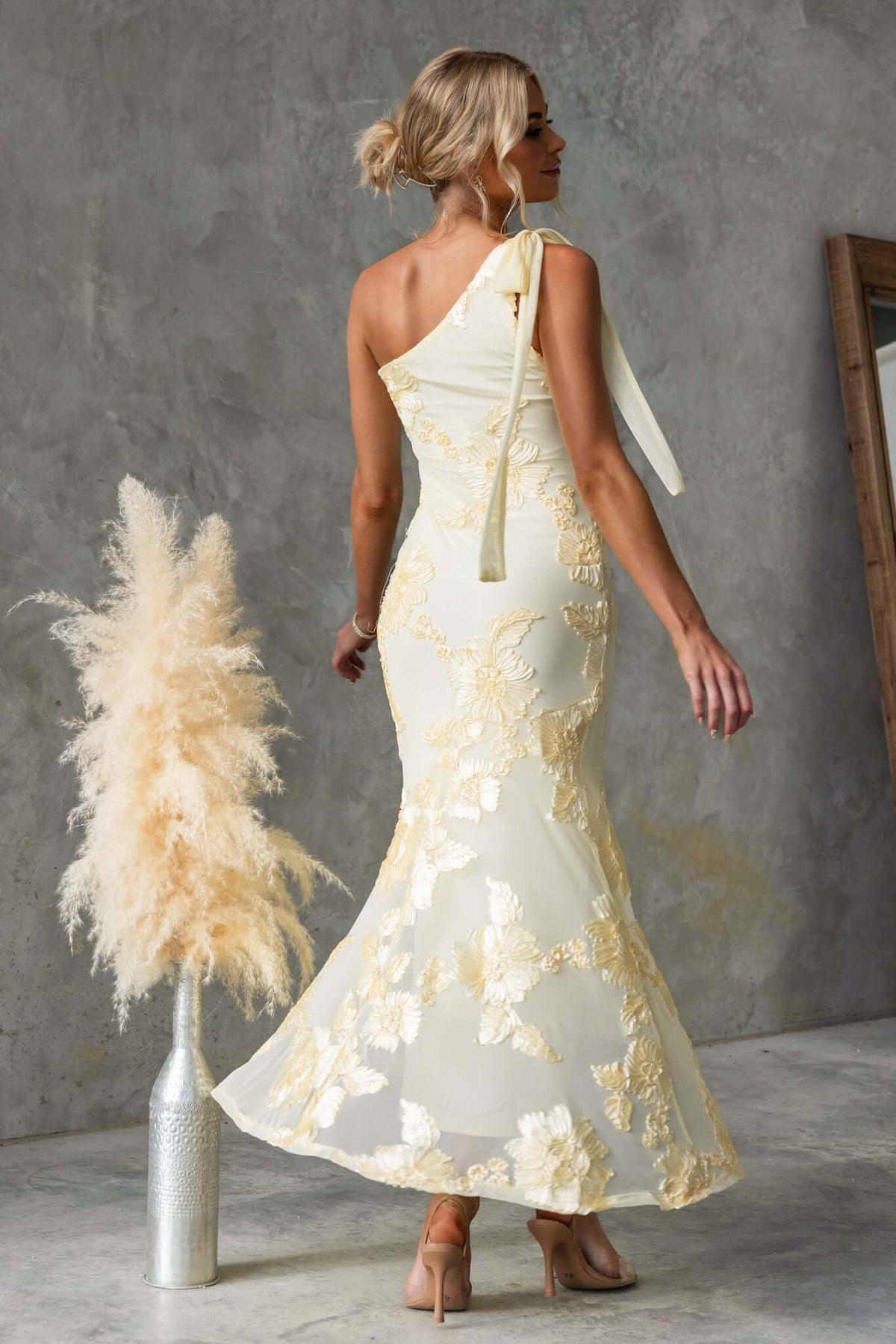 Inspire Maxi Dress in yellow embroidered fabric, featuring a one-shoulder design and side zip, styled with a vase and pampas grass.