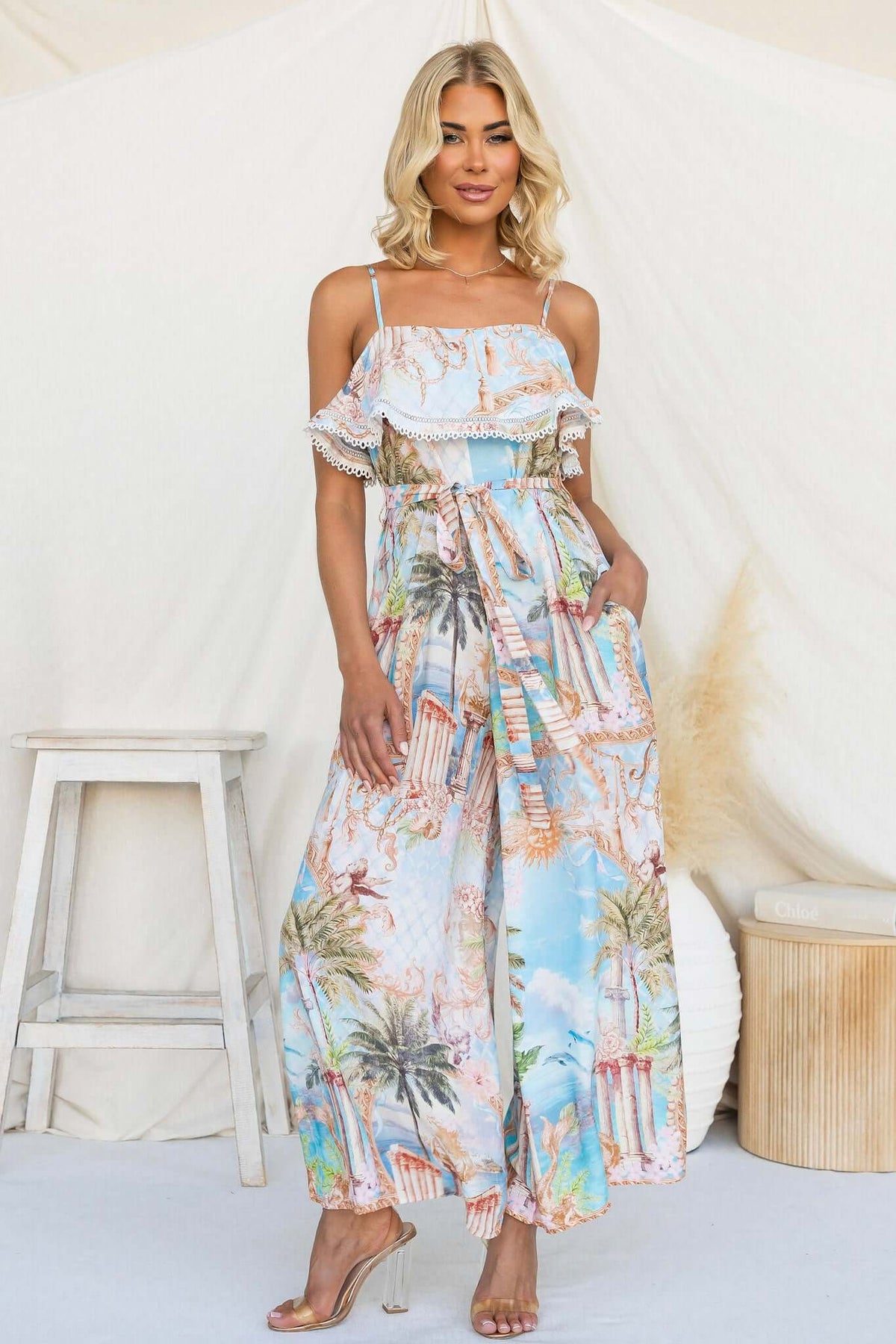 Model wearing Island Escape Jumpsuit with adjustable straps and floral print in a bright beach setting.