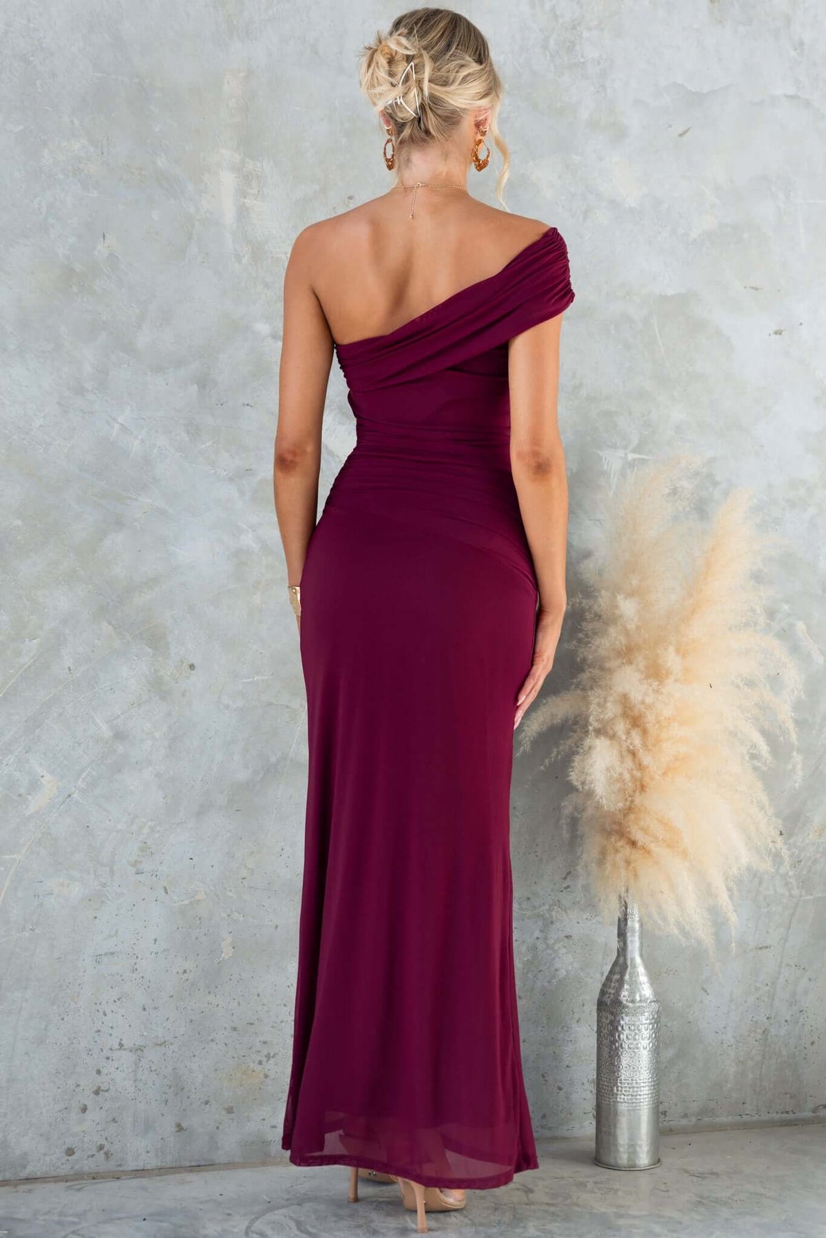 Back view of a woman in a burgundy Korren Maxi Dress with one-shoulder neckline and ruched design.