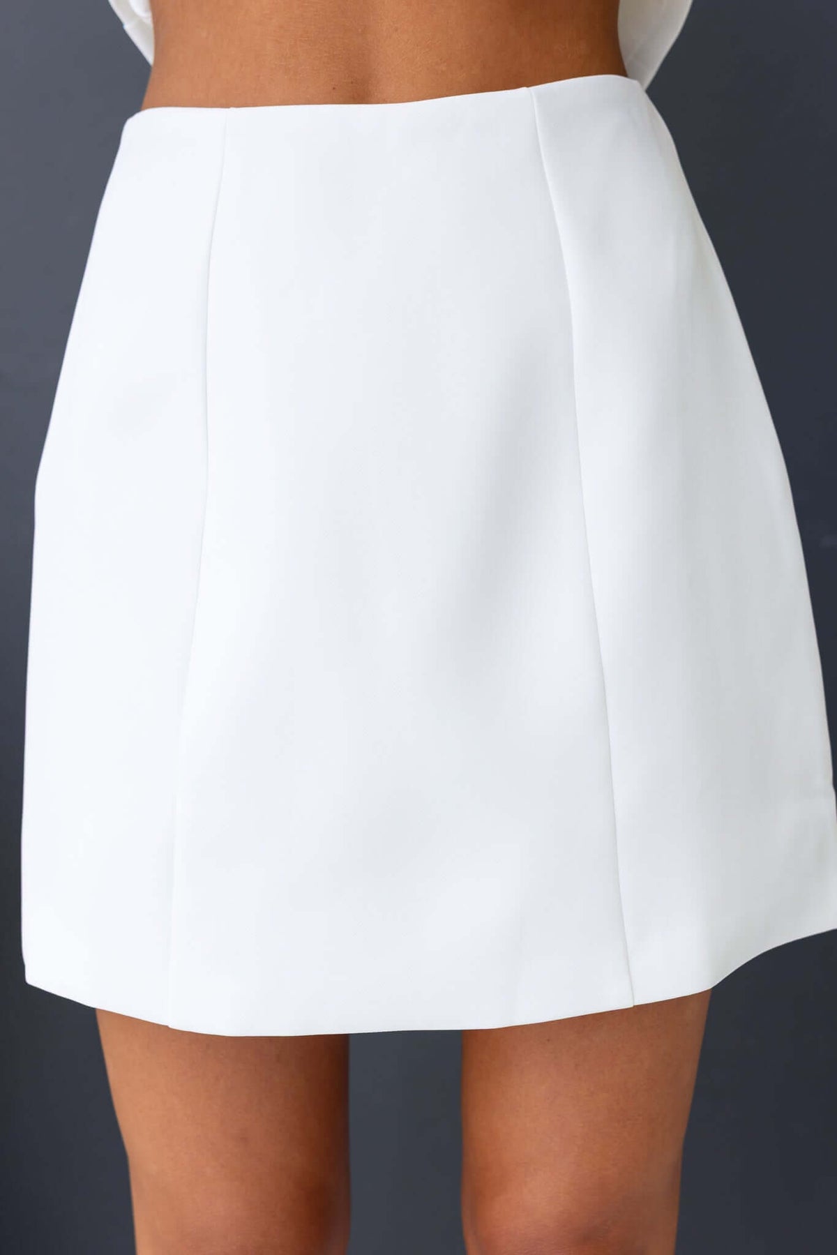A model showcases the Laredo Skirt, a mid-high waist white skirt with a clean design and invisible zip detail at the back.