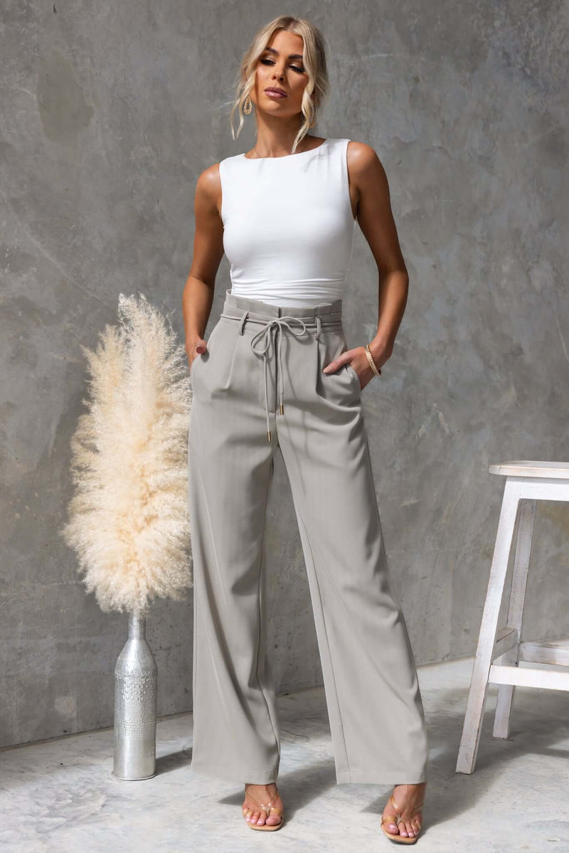 Lesky Pants in light grey, featuring a paperbag waist, removable tie, and pockets, modeled by a woman in a stylish setting.