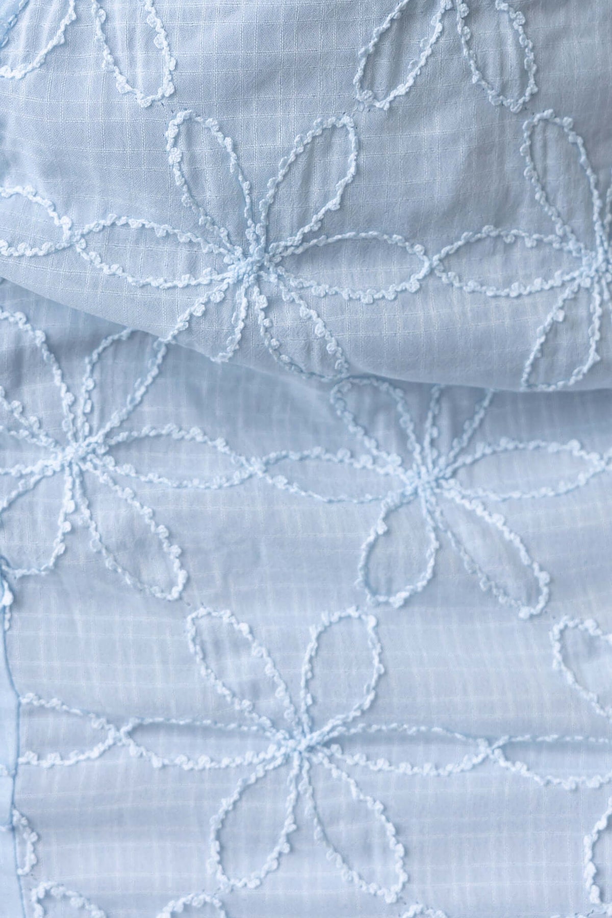 Close-up of light blue textured fabric with embroidered floral pattern, featuring petals and intricate details.