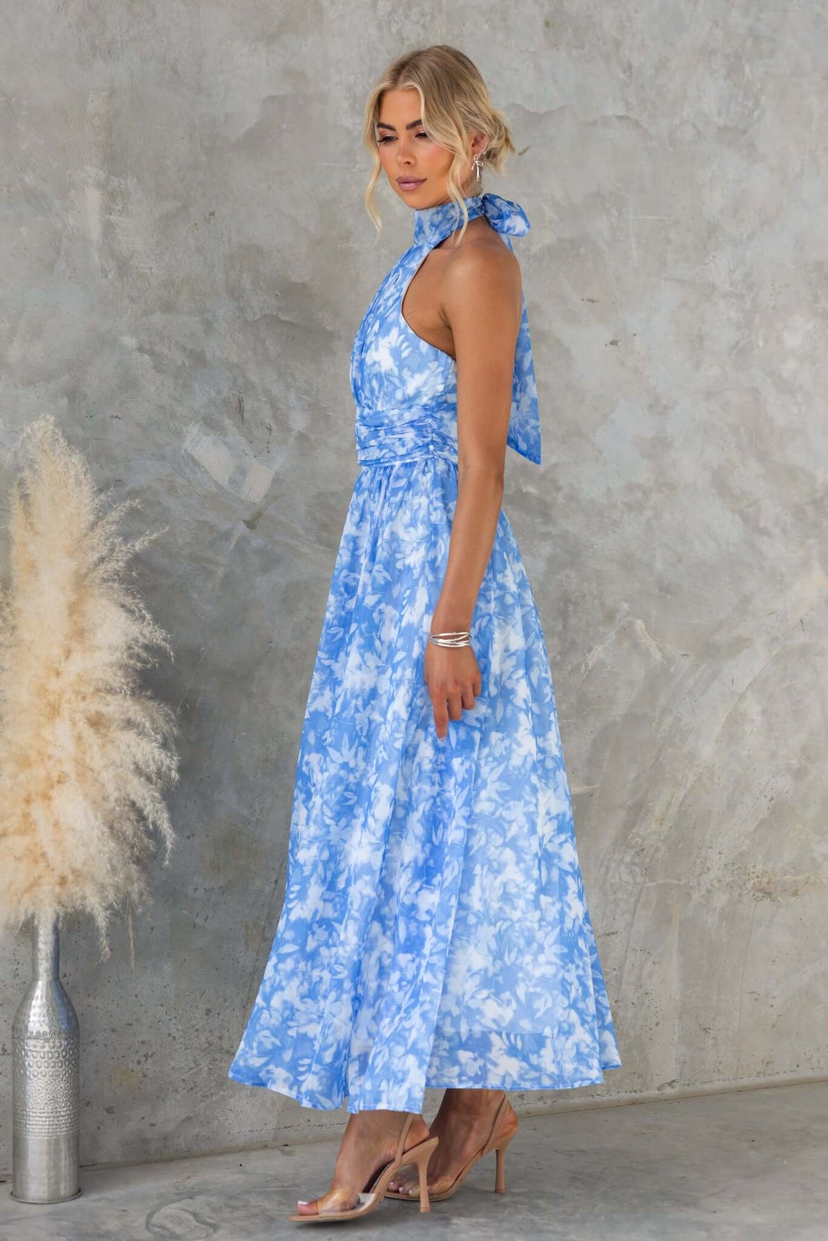 Model wearing a blue floral Little Things Maxi Dress with a high tie neckline against a textured wall.
