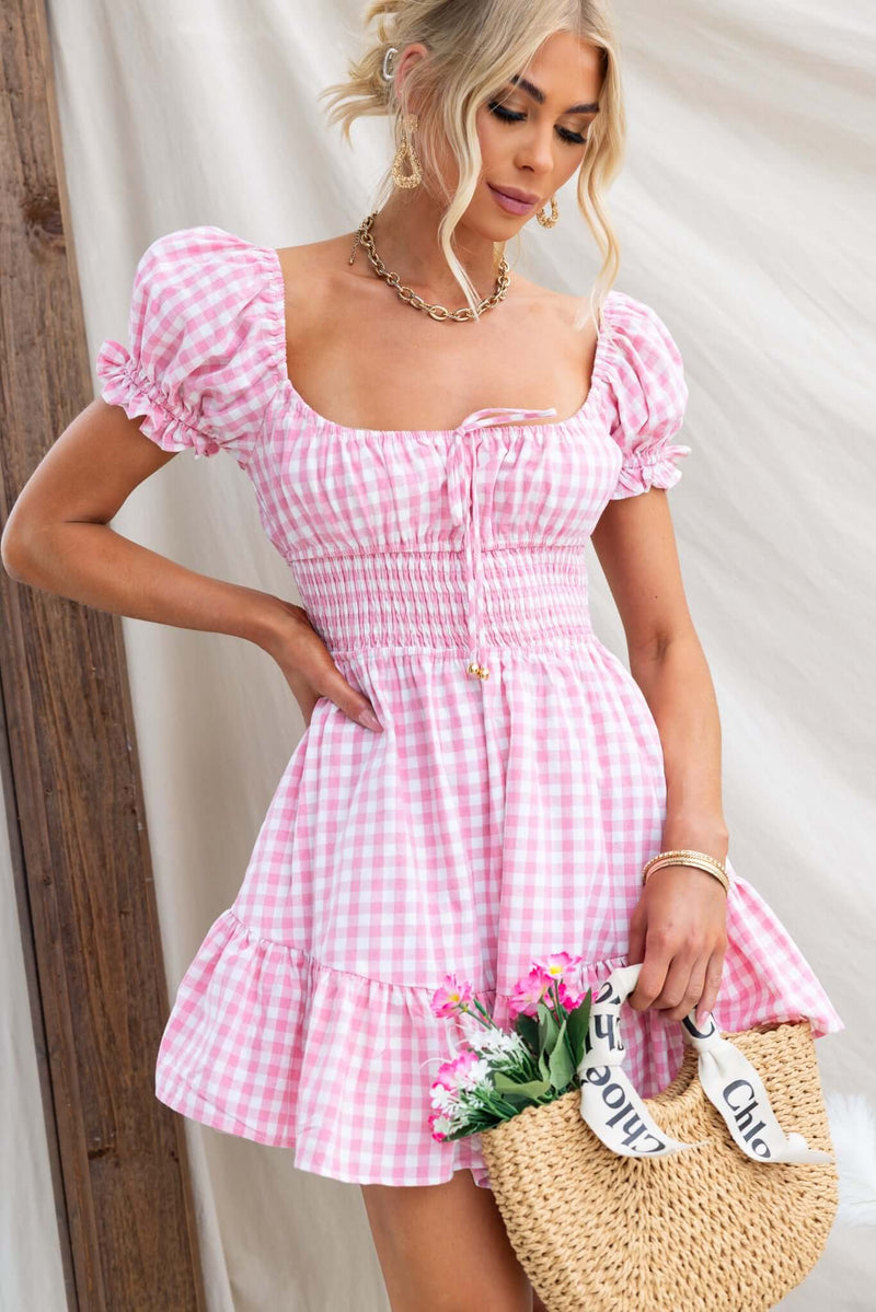 Model wearing a pink gingham Love In Motion Dress with puff sleeves and a flared skirt, holding a straw bag and flowers.
