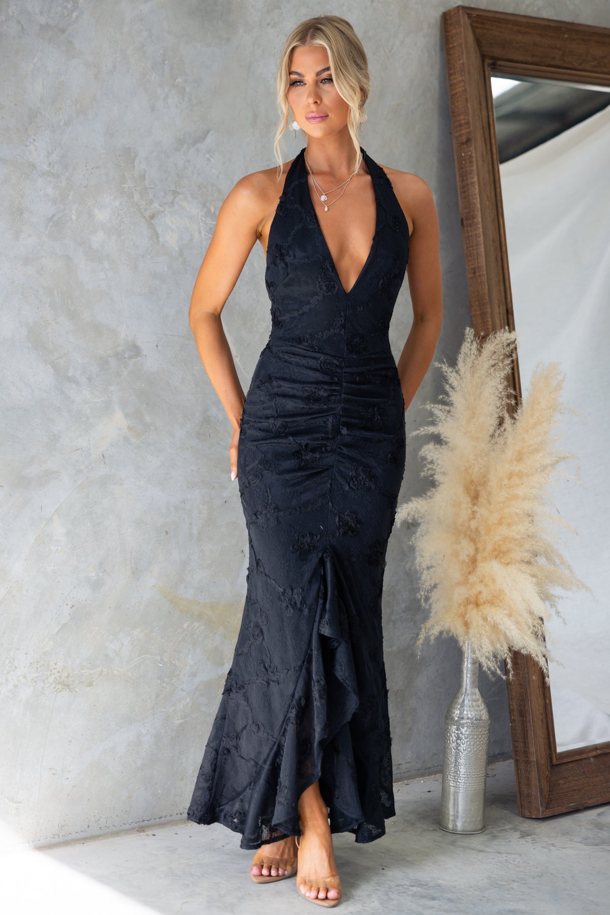 Elegant black Metrick Maxi Dress with halter V-neckline, textured material, and fishtail hem, model showcasing it in front of a mirror.
