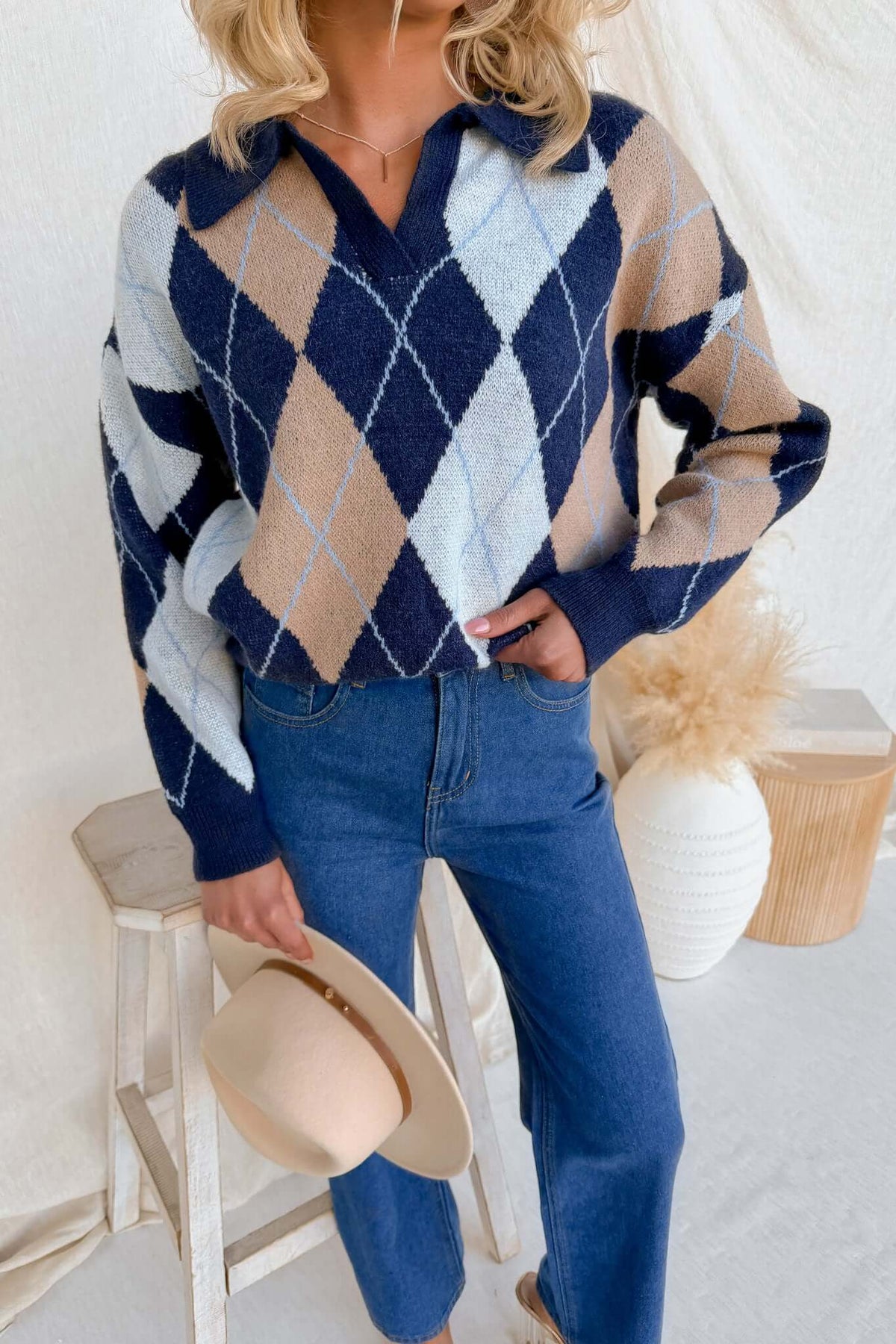 Model wearing Vella Jumper in navy diamond pattern, holding a hat while styled with denim jeans.