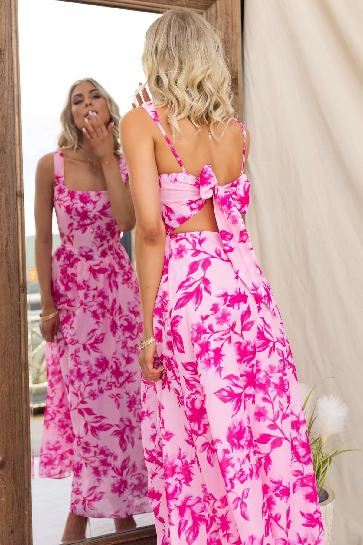 Okansa Maxi Dress in pink floral print with adjustable straps and tie back, showcased by model in front of a mirror.