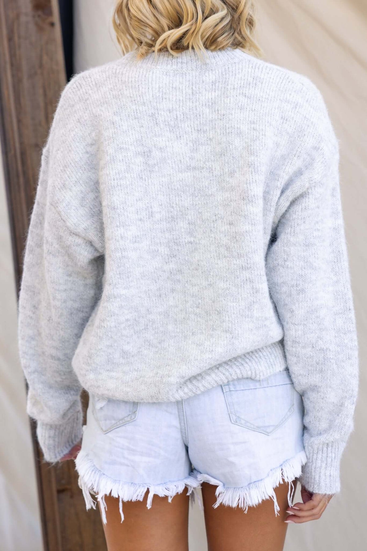 Back view of a model wearing a gray Palmyra jumper with round neckline and relaxed fit, paired with distressed denim shorts.