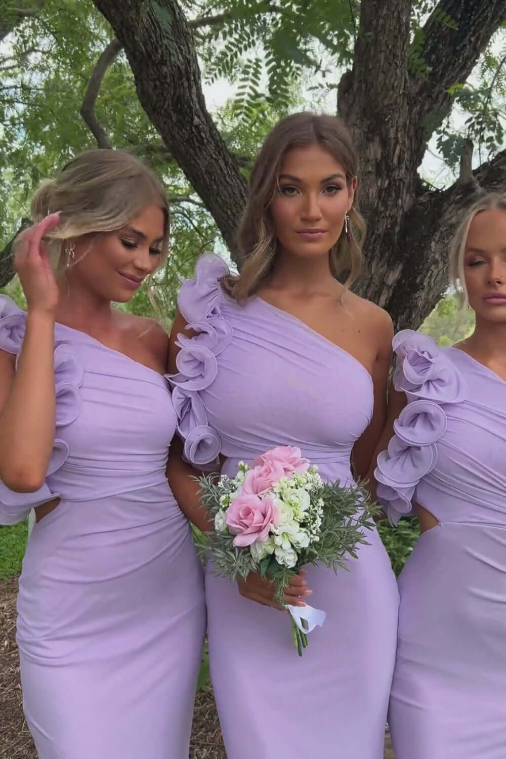 Three models in lilac Meriden Maxi Dresses with one-shoulder necklines and ruffled sleeves, holding bouquets.