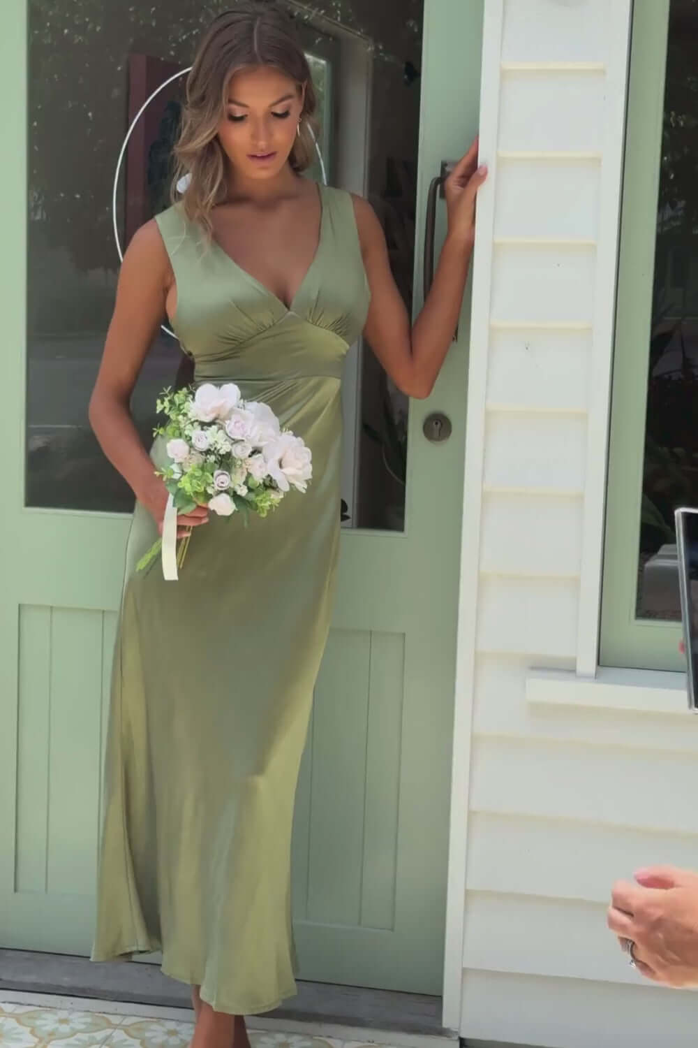 Model wearing Alika Maxi Dress in olive green, V-neckline, holding bouquet, standing by a door.