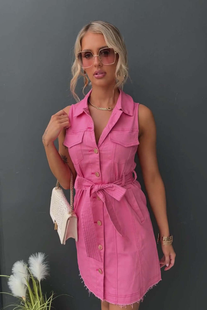 Model in a pink denim dress with button-up front and tie waist, accessorized with sunglasses and a handbag.