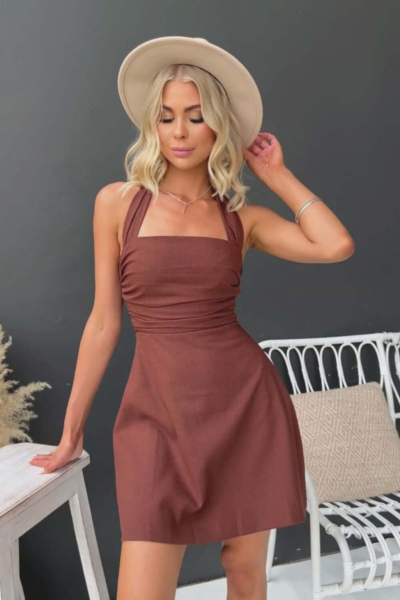 Quade Dress featuring tie halter neckline, back zip closure, and a rich brown color, styled with a fashionable hat.