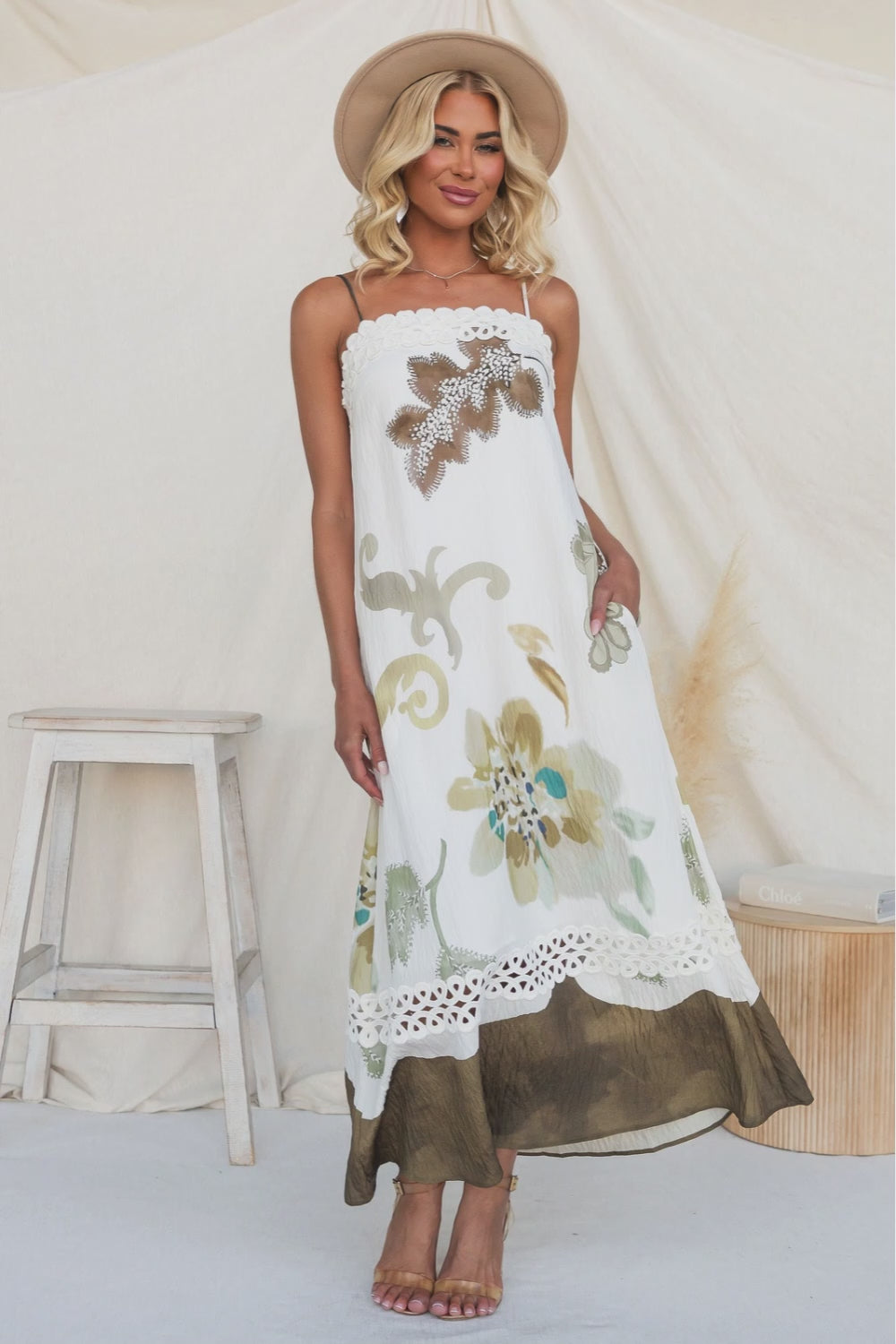 Model wearing Carlo Maxi Dress with floral design and tie straps, standing by a wooden stool.