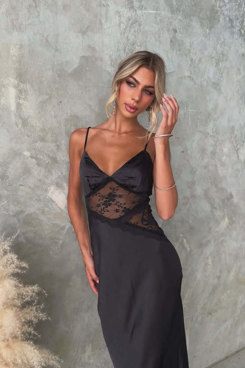 Model in an elegant black Obsidian Maxi Dress with lace panel and adjustable straps against a textured wall.