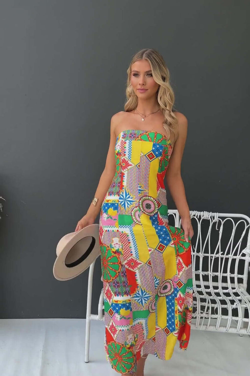 Woman wearing a colorful strapless Infinity Maxi Dress with vibrant patterns, holding a hat and posing indoors.